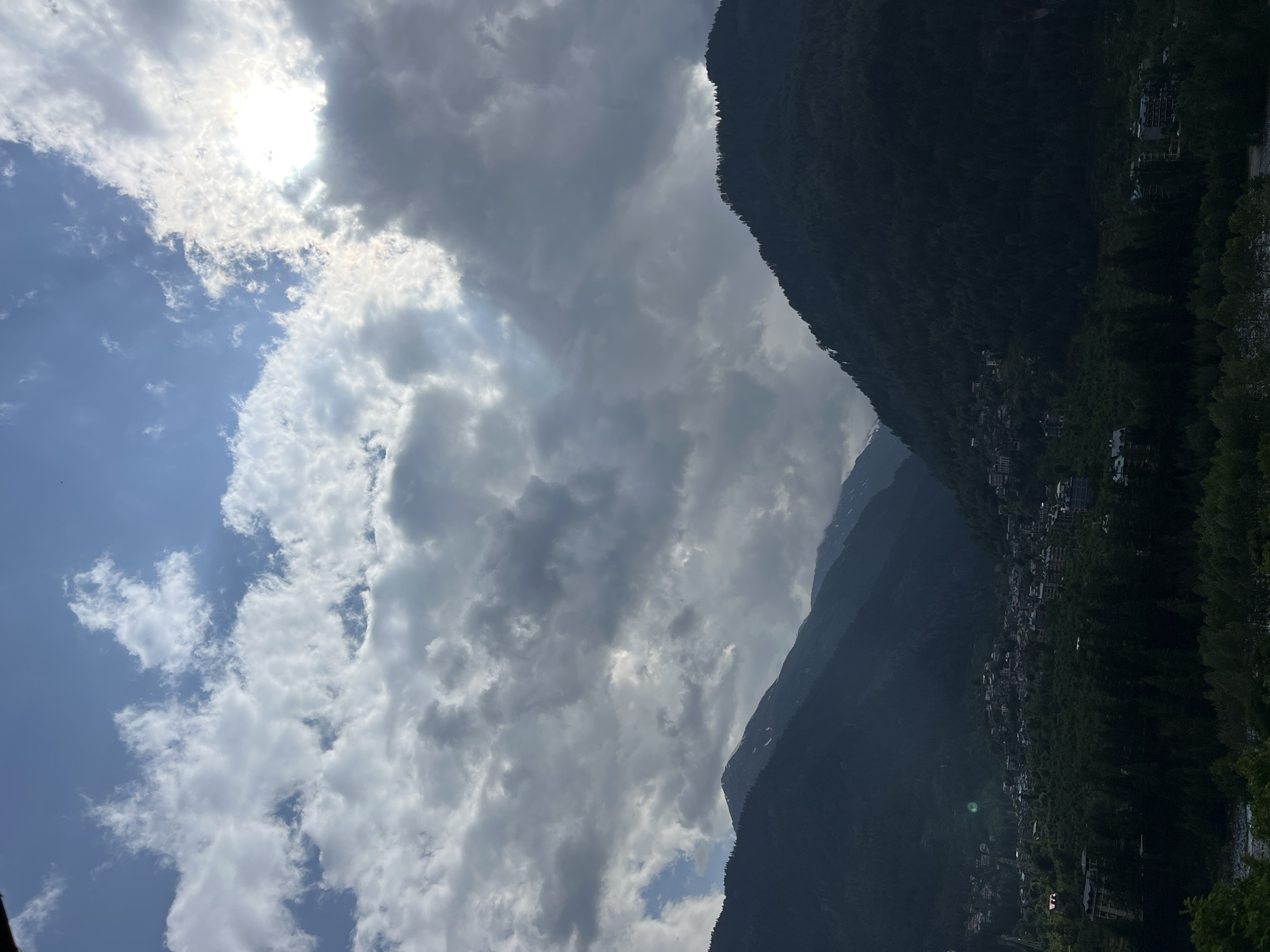 The image captures a picturesque mountain landscape in Vashisht, India, under a dramatic, cloudy sky. The scene is dominated by imposing, dark mountains, heavily forested with dense trees, receding into the distance. A village or town is nestled along the slopes and base of these mountains, featuring numerous buildings with varying architectural styles, suggesting a settled community within the rugged terrain.
The sky occupies a significant portion of the frame, presenting a dynamic display of weather conditions. Bright sunlight pierces through thick, white clouds in the upper left, creating a radiant glow. Adjacent to this, large, grey, and darker clouds stretch across the sky, casting shadows and giving a sense of depth and impending weather. Patches of clear, light blue sky are visible in between the cloud formations, adding contrast. The overall lighting suggests a time of day when the sun is relatively high, possibly late morning or early afternoon, but largely obscured by the heavy cloud cover.
No specific human activity is discernible, and there is no visible text. The image evokes a sense of tranquil grandeur and the coexistence of human habitation with vast natural beauty, characteristic of a Himalayan valley.
