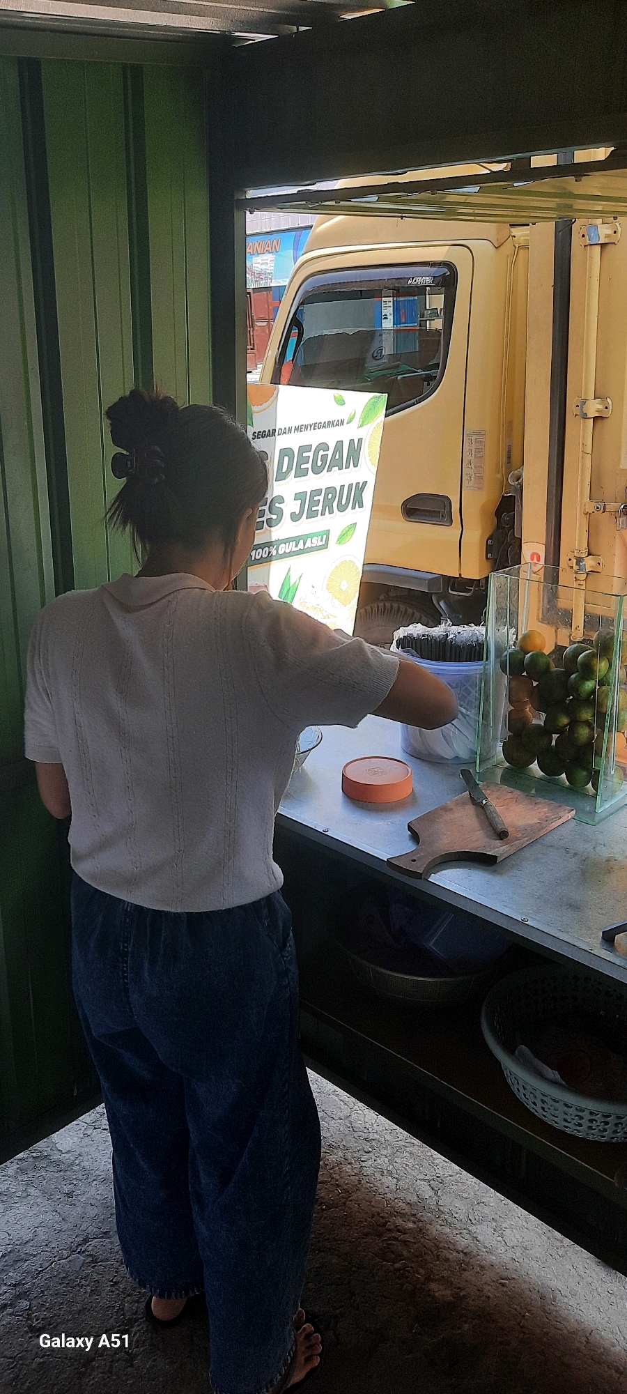 In Selopuro, Indonesia, a woman stands behind a counter, facing away from the camera. She is wearing a white, short-sleeved collared shirt and dark blue wide-leg pants. Her hair is pulled up in a bun with a dark clip. In front of her, on the counter, is a bright orange lid and a small wooden cutting board with a knife. To her right, a glass display case contains a stack of green citrus fruits, likely oranges or limes. Behind the woman, a large, yellow truck is visible, its side door open. A prominent sign attached to the truck advertises "DEGAN ES JERUK" (Coconut Ice Orange), with the text "SEGAR DAN MENYEGARKAN" (Fresh and Refreshing) and "100% GULA ASLI" (100% Real Sugar). The background suggests an outdoor setting with a corrugated green wall on the left and the truck's cargo area on the right. The ground is paved with an uneven, textured surface.