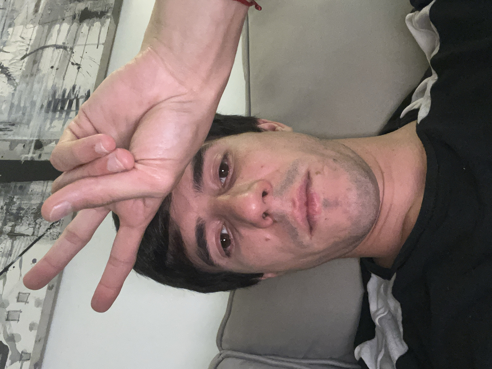 A casual portrait captures a single male lying down indoors, likely in Buenos Aires, Argentina. The image is oriented with the subject's head to the right and his body extending left, suggesting it might be a rotated selfie. He rests his head on a plain grey fabric pillow or cushion.
The man has dark hair and dark eyes, looking directly at the camera with a neutral to slightly contemplative expression. He has short stubble on his face and a small mole visible below his right eye. His left hand is raised above his head, forming a "peace" or "victory" sign with his index and middle fingers extended. A thin red bracelet adorns his left wrist. The skin on his hand shows visible veins and creases, and the fingernails are short and unpolished.
He is wearing a black t-shirt featuring a white, possibly ruffled or folded, fabric design on the shoulder and sleeve area. In the background, behind his head and hand, an abstract artwork is visible on the wall. This piece displays a dynamic pattern of black and white splatters, drips, and brushstrokes, suggesting a modern artistic style. The lighting appears soft and internal, typical of an indoor setting. No discernible text is visible in the image.