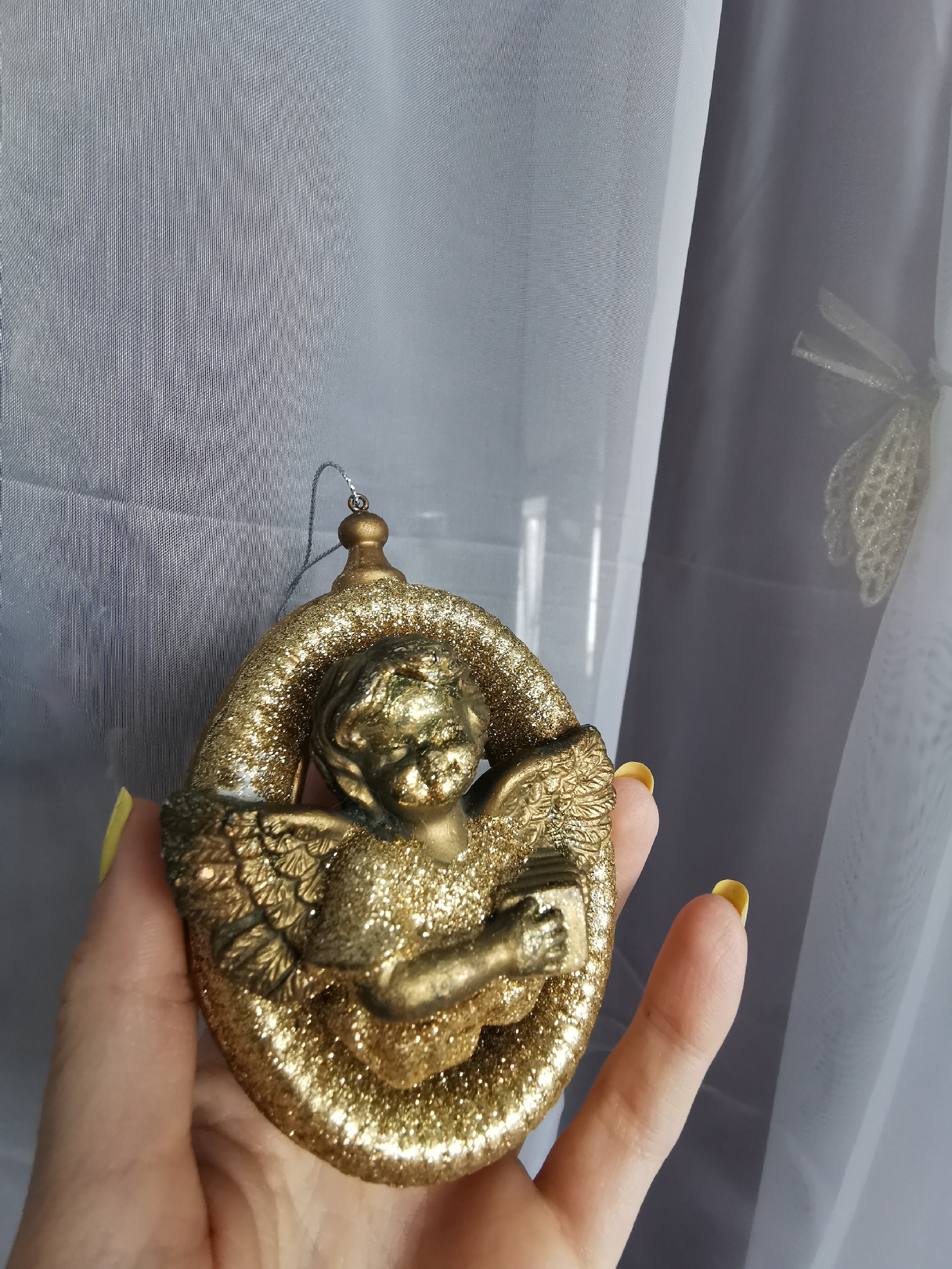 A person is holding a gold-colored angel ornament in their hand. The ornament has a glittery finish and features an angel with wings, holding a book. The background consists of sheer white and grey curtains. The location is not specified.