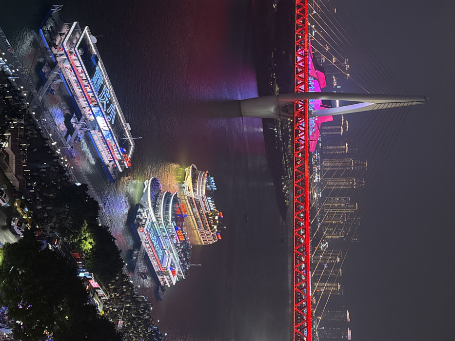A high-angle nighttime photograph depicts an illuminated riverfront scene in Yuzhong District, Chongqing, China. The Dongshuimen Bridge dominates the mid-ground, its main structure and cables aglow in red light, with its central cable-stayed pylon illuminated in white and purple. Beyond the bridge, a dense cityscape of high-rise buildings is visible, outlined by numerous yellow and white lights. Directly beneath the bridge's red span, a large, distinctive building complex is illuminated in vibrant pink and red hues.
On the river below, three multi-deck passenger cruise ships are present. The ship furthest upstream is brightly lit with white, gold, and red lights. Closer to the foreground, a second ship, adorned with white, blue, and red illuminations, is either docked or moored. In the immediate foreground, a third, elongated multi-deck ship is prominently docked, displaying blue, white, and red lights, including large blue characters on its upper deck.
The riverbank in the foreground is a bustling promenade crowded with a large congregation of people. These individuals are densely packed, particularly around the docking areas, where multiple gangways connect to the vessels. Adjacent to the promenade, an urban street is visible, with several vehicles, including cars and at least one bus, indicating vehicular traffic. Scattered streetlights illuminate the immediate vicinity of the promenade and street. The dark river surface reflects the colorful artificial lights from the bridge, buildings, and boats.