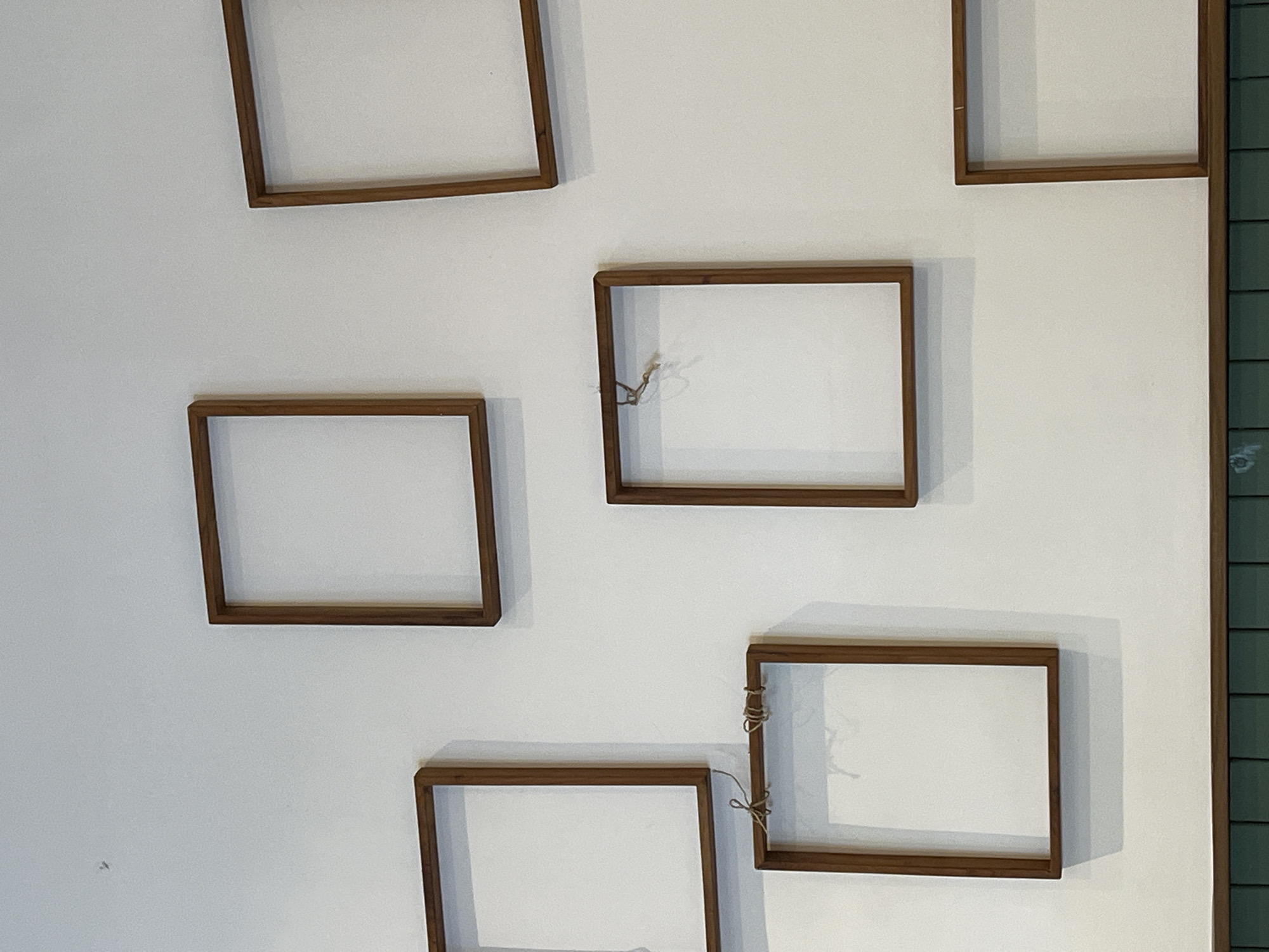 A close-up shot shows a section of a white wall adorned with multiple empty rectangular wooden frames. Some frames have remnants of twine or string attached. To the right of the frame is a section of what appears to be vertically stacked, dark-greenish tiles. The location is likely in Kuta Selatan, Indonesia, though this cannot be definitively determined from the image itself. No people or actions are discernible.