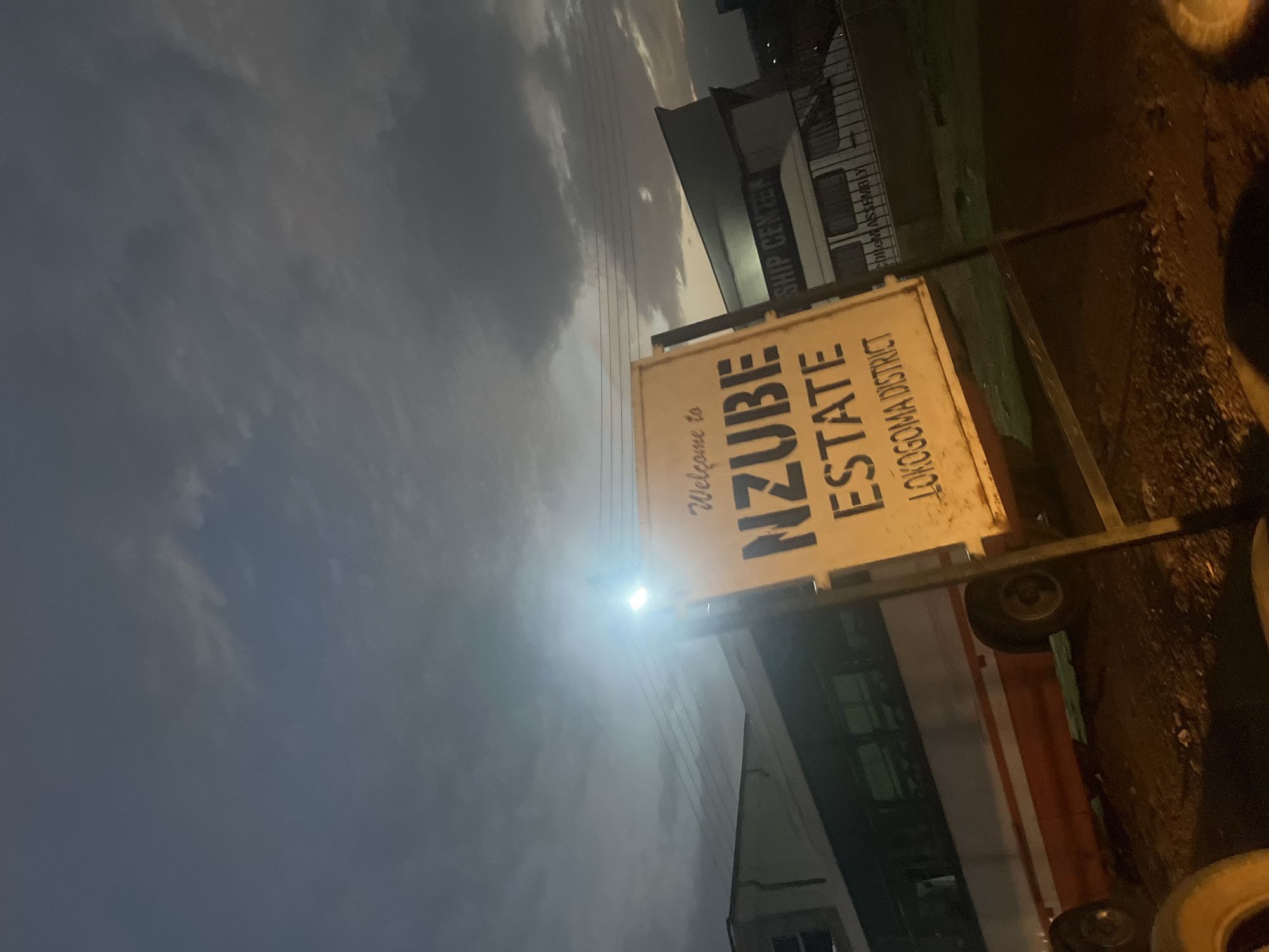 This image captures an outdoor scene at dusk or early night, characterized by low light and an overcast sky. The primary subject is a brightly illuminated, rectangular sign positioned centrally in the frame. The sign reads, "Welcome to NZUBE ESTATE LOKOGOMA DISTRICT." It appears to be a freestanding sign, possibly on a wheeled frame.
In the background, several buildings are visible. One prominent structure features text that includes "SHIP CENTER" and partially "MASS ASSEMBLY" (or similar). Overhead, power lines stretch across the dark, cloudy sky, typical of an urban or semi-urban area. A strong artificial light source, possibly a streetlamp or part of the sign's illumination, creates a significant glare near the top of the sign. The ground in the foreground is dark and indistinct, suggesting an unpaved surface, with some shadowy objects resembling tires or debris.
The visible text explicitly identifies the location as "NZUBE ESTATE" within the "LOKOGOMA DISTRICT," which is a known area in Abuja, Nigeria, aligning with the provided context. The overall atmosphere is one of transition, between day and night, with artificial lights beginning to dominate. No people or specific activities are visible.