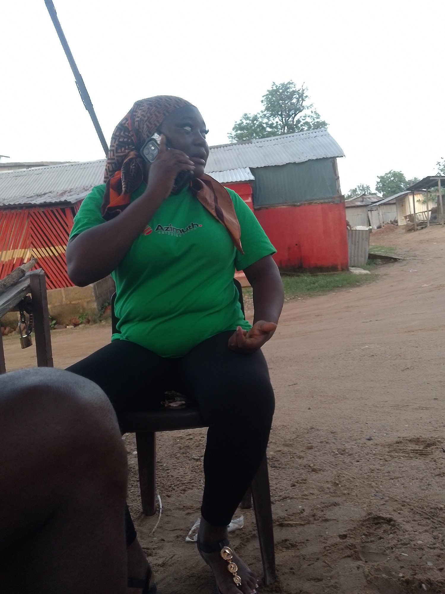 The image captures a candid outdoor scene in what appears to be Adankolo, Nigeria, featuring a person engaged in a phone call. The main subject, a dark-skinned individual, is seated on a brown plastic chair, looking to the right while holding a smartphone to their left ear. They are wearing a green short-sleeved t-shirt with a logo that reads "Azimuth." (with another word, possibly "outside," partially obscured below it), black pants, and open-toed sandals with gold-colored decorative elements. A patterned brown and orange headscarf covers their head. Part of another person's leg or lap is visible in the immediate foreground on the bottom left.
The setting is an unpaved, dusty area, likely a dirt road or yard. In the background, several simple buildings with corrugated metal roofs are visible, painted in colors like red and green, suggesting a semi-urban or village environment. One building has distinctive red and black diagonal stripes. A wooden table-like structure with a padlock attached is next to the seated person. The sky is bright and appears hazy or overcast, indicating daytime, possibly mid-morning or afternoon, given the soft lighting. Scattered trees are visible further in the background. The overall atmosphere suggests a casual, everyday moment.