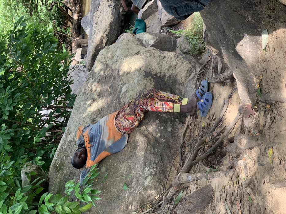 A man is lying on his back on a large rock, propped up on his elbows, in Egbeda, Nigeria (latitude: 7.119, longitude: 5.362). He is wearing a long-sleeved, orange and gray shirt with a faded pattern and colorful patterned pants. His bare feet are wearing blue sandals. There is a large, gray rock behind him, along with a small blue bucket to the right of the frame. The man's eyes are closed and he appears to be sleeping or resting. There is dense foliage and vegetation in the background, as well as a small stream. The scene is partially shaded from the sun by trees. The ground is sandy and there are large roots protruding from the soil, likely from the nearby trees.