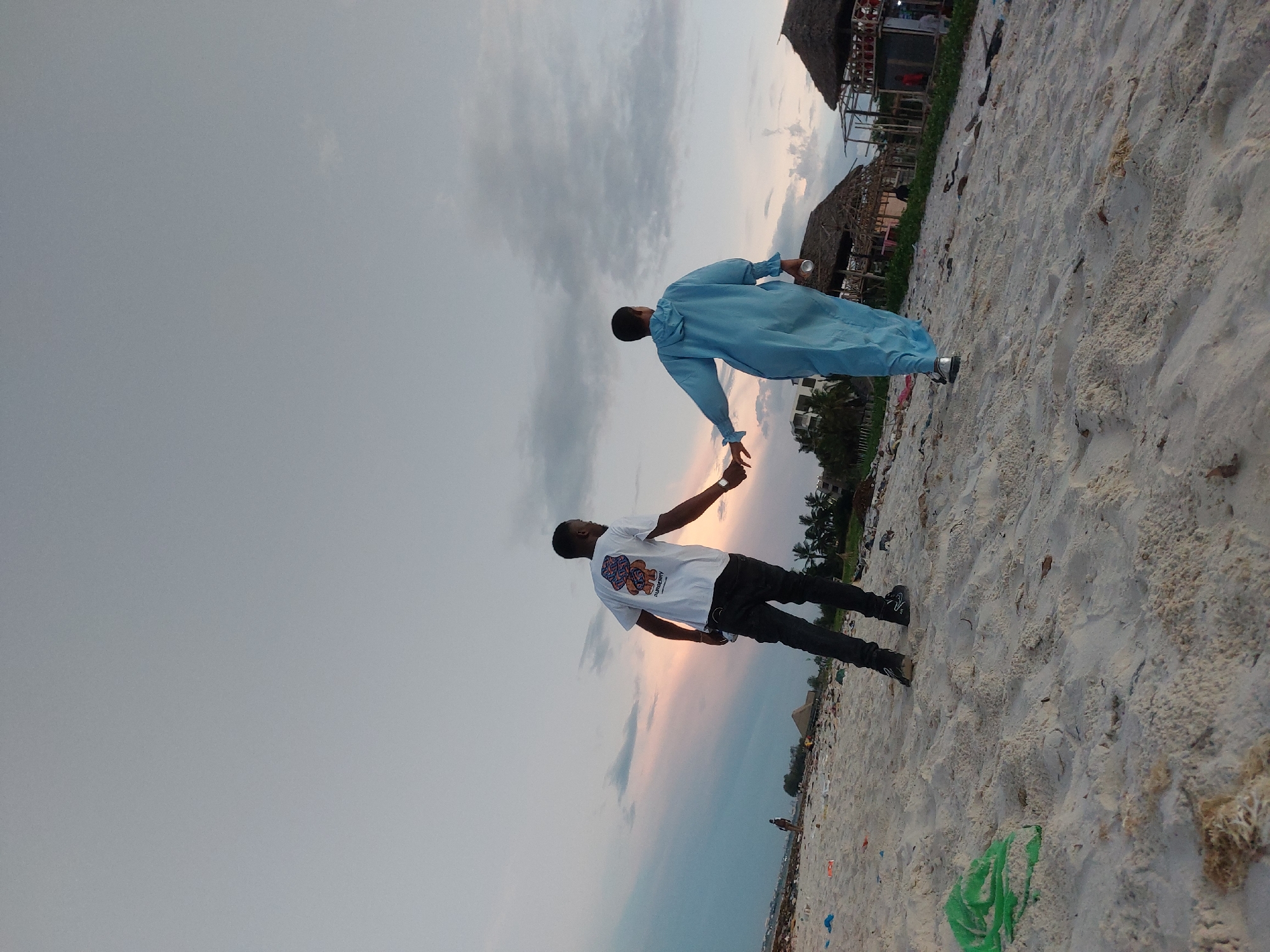 The photo depicts a scene on a beach in Dar es-Salaam, Tanzania. Two young men are interacting. One man, wearing a white t-shirt and dark pants, reaches out and touches the hand of another man, who is dressed in a light blue robe-like garment. They appear to be standing on a sandy beach littered with debris and trash. In the background, there are buildings with thatched roofs and palm trees. The sky is overcast with a pinkish hue near the horizon, indicating either sunrise or sunset.