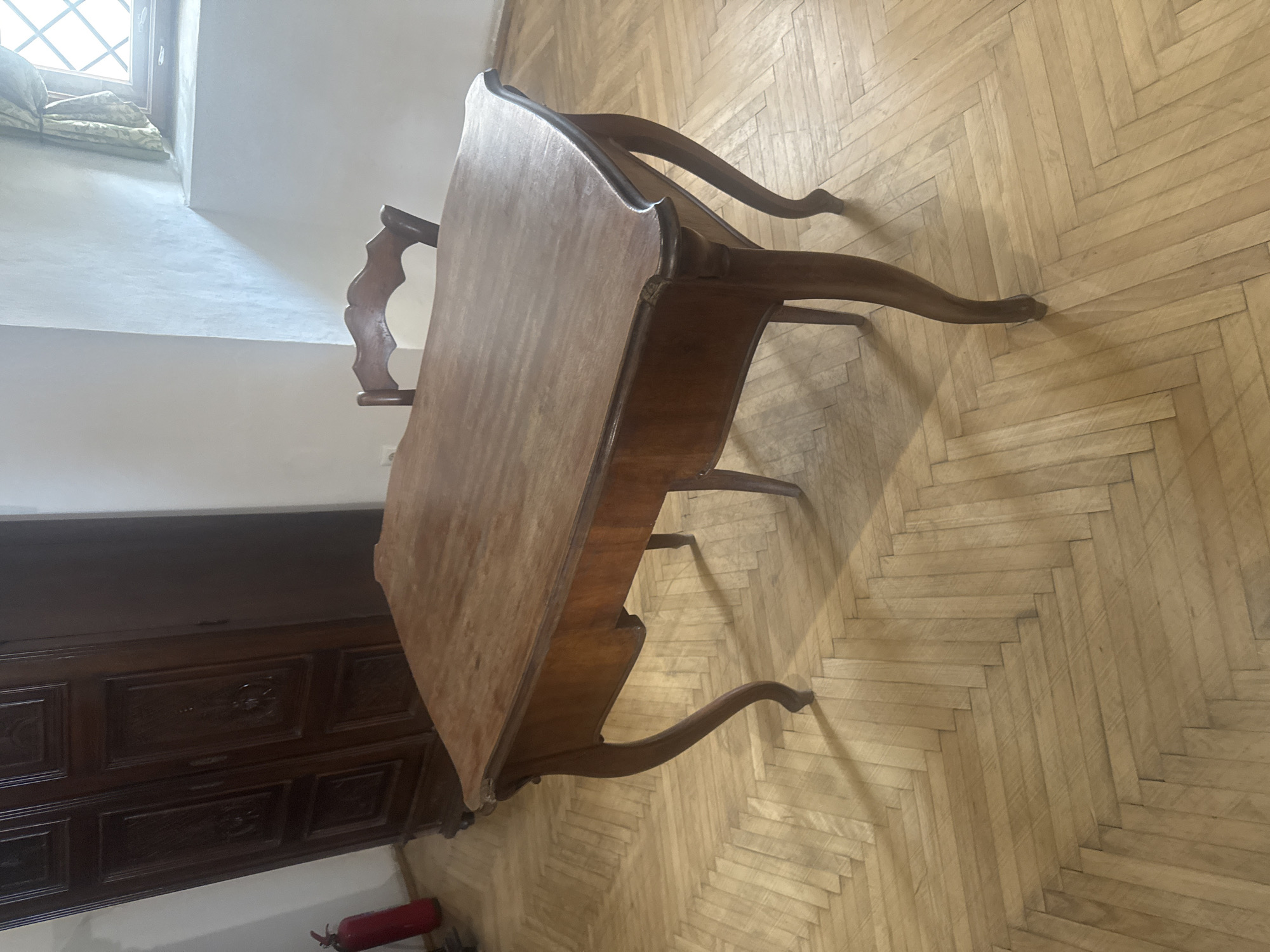 A high-angle shot depicts a wooden desk in a room, positioned at an angle against a herringbone-patterned wooden floor. The desk, made of dark wood, has ornate carved details on its sides and legs. A chair with a similarly carved back is partially visible behind the desk. To the left of the desk, a large, dark-wood cabinet or dresser is seen with carved floral motifs on its drawers and panels. In the lower left corner, a red fire extinguisher is positioned near the base of the cabinet. The room is lit by natural light coming from a window in the upper left corner, casting shadows across the floor.