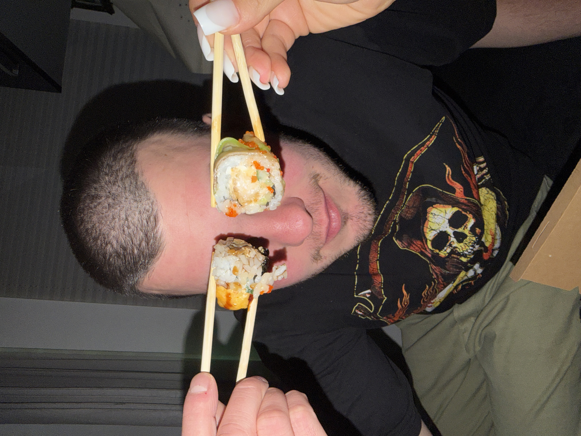 A fair-skinned man is photographed from a tilted perspective, facing the viewer. Two pieces of sushi are held up to his eyes with wooden chopsticks. The sushi is cylindrical, with rice, some type of filling, and orange roe. He is wearing a black long-sleeved shirt with a skull and crossbones graphic. The environment appears to be indoors. There is no information to determine if the image was taken in Varna, Bulgaria.