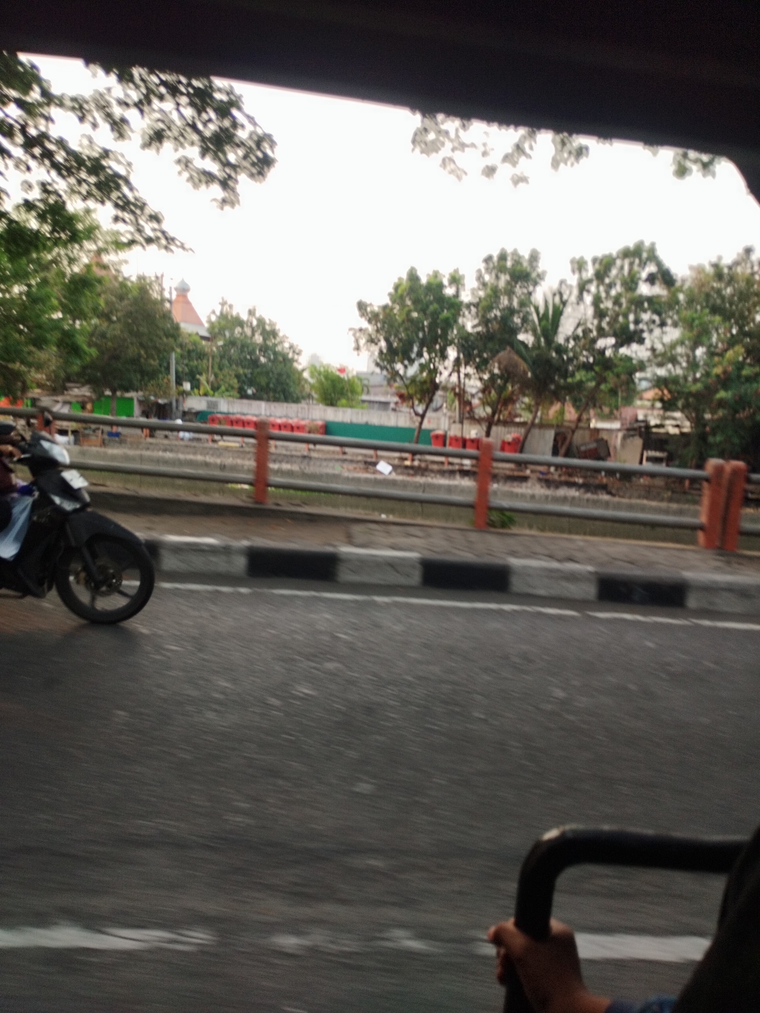 The image shows a blurry, motion-streaked view of a street scene, likely from inside a vehicle. On the left, a black motorcycle is partially visible, with a person's arm, wearing blue, gripping its handle. The motorcycle is in motion. The background features a road with a gray barrier and reddish-orange posts. Beyond the barrier, there are some buildings and trees. The sky is bright and overcast. The overall impression is that the photo was taken while moving at a moderate speed. The presence of the motorcycle, urban infrastructure, and vegetation suggests a location within a city.