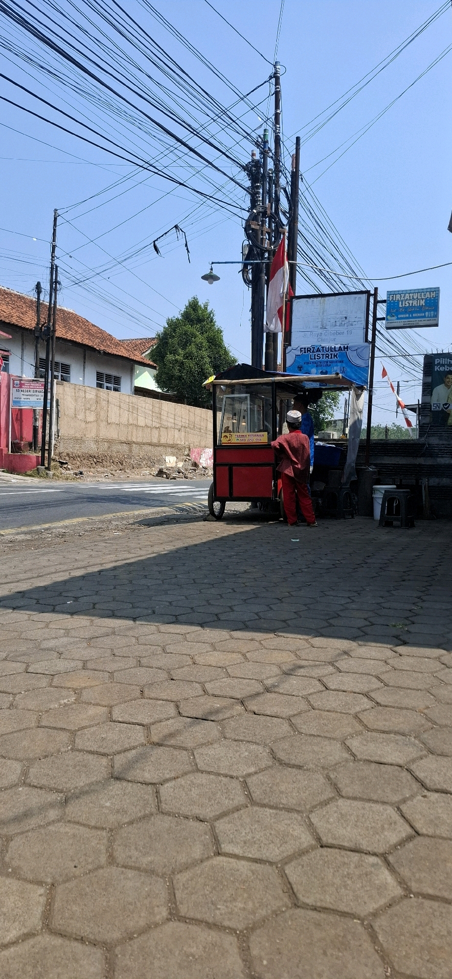 The image shows a street scene in Sirnagalih, Indonesia. In the foreground, a hexagonal brick sidewalk is visible, with a shadow cast upon it from the right. A red food cart, with a glass display case and metal framing, is situated on the side of the road. A person wearing a red uniform and a white cap is standing behind the cart, interacting with another person inside. The cart has a sign in yellow that reads "TERIMA PESANAN" (Take Orders). A white and red Indonesian flag is displayed on a pole behind the cart. Beyond the cart, a street runs horizontally, with a pedestrian crossing marked by white stripes. A low concrete wall borders the street, behind which are buildings with red tiled roofs and white walls, and a large green tree. Overhead, a dense network of electrical wires crisscrosses the frame against a clear blue sky. Two signs are visible on poles near the cart: one larger sign for "FIRZATULLAH LISTRIK" (Firzatullah Electricity) and a smaller blue sign with similar text. The scene appears to be a typical roadside business operating in a residential or commercial area.