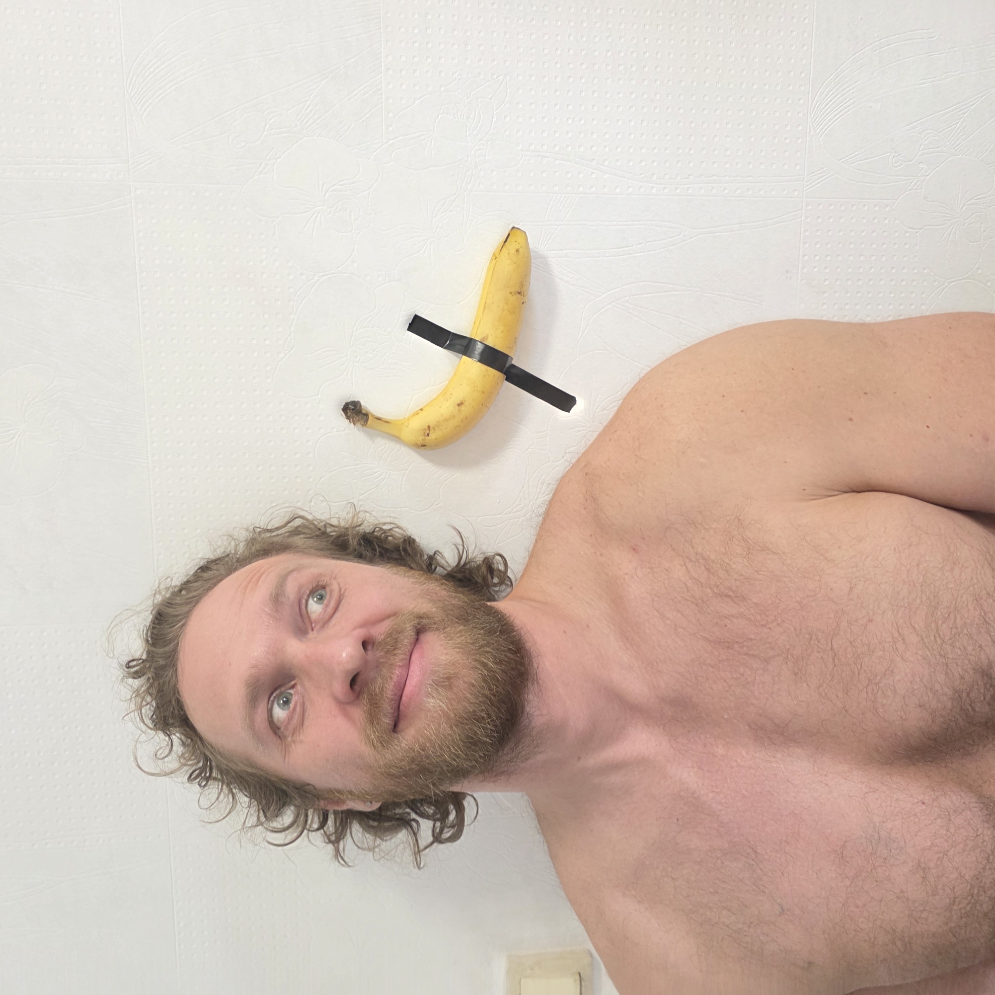 A man with a beard and curly hair is looking upwards. A banana is taped to a white textured wall above him. The man is shirtless. A light switch plate is visible in the lower right corner. The specific location in Saint Petersburg, Russia is not evident from the image.