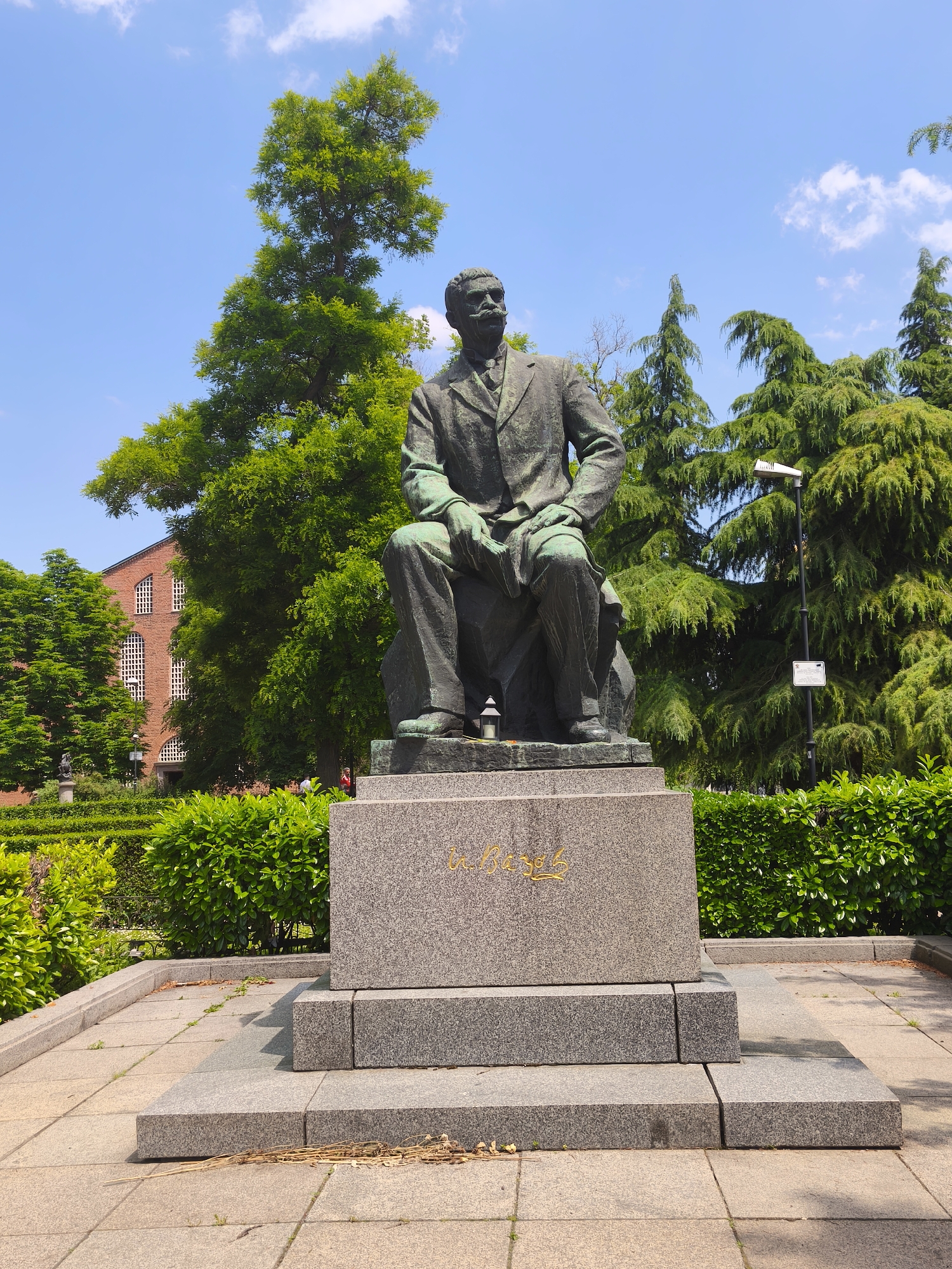 A full-frame, eye-level outdoor photograph captures a bronze statue of a seated male figure, Dr. Hristov, on a multi-tiered granite pedestal, located in the Doctor's Garden (Doktorska Gradina) in Sofia, Bulgaria, under a bright blue sky with scattered white clouds.
The statue, depicting a man with a prominent mustache wearing a suit, appears patinated with green oxidation, indicative of bronze. The figure is seated on a rough-hewn stone bench, which is part of the statue itself, with hands resting on his lap. A small, dark decorative lantern or candle holder is placed directly on the statue's base near the figure's feet. The inscription "Д-р Христов" (Dr. Hristov) in golden script is visible on the front face of the main, light-gray granite plinth. The pedestal consists of several rectangular granite blocks, forming three steps leading up to the main plinth.
The monument is centered on a paved area composed of square, light-colored concrete slabs. Scattered dry leaves, twigs, and small patches of weeds are visible on the paving around the base of the monument. Surrounding the paved area are neatly trimmed, vibrant green bushes forming a low hedge.
In the background, a variety of trees are prominent. To the left of the statue, a tall deciduous tree with bright green foliage reaches towards the sky. Further left, a large, multi-story building constructed of red brick with distinctive arched white-framed windows is partially visible behind the trees. A faint human figure in a red top is discernible near this building. To the right of the statue, several dense, dark green coniferous trees are present. A tall, black lamppost with a modern rectangular light fixture and a small white informational sign attached to it stands between the statue and the coniferous trees on the right. Bright sunlight illuminates the scene, casting soft shadows on the ground and the monument.
