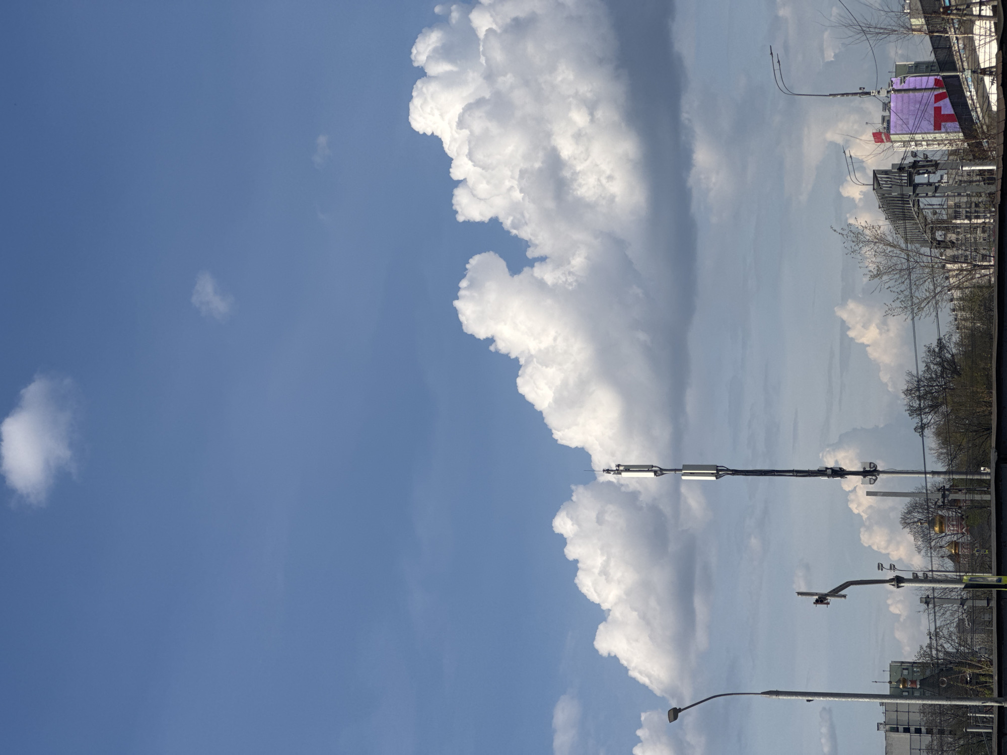The image shows a daytime view in Moscow, Russia, with a blue sky and large cumulus clouds dominating the upper portion. Power lines and light poles are visible in the lower portion, along with bare trees and buildings. A partially visible advertisement with a red cross is seen on a building towards the right side of the frame.