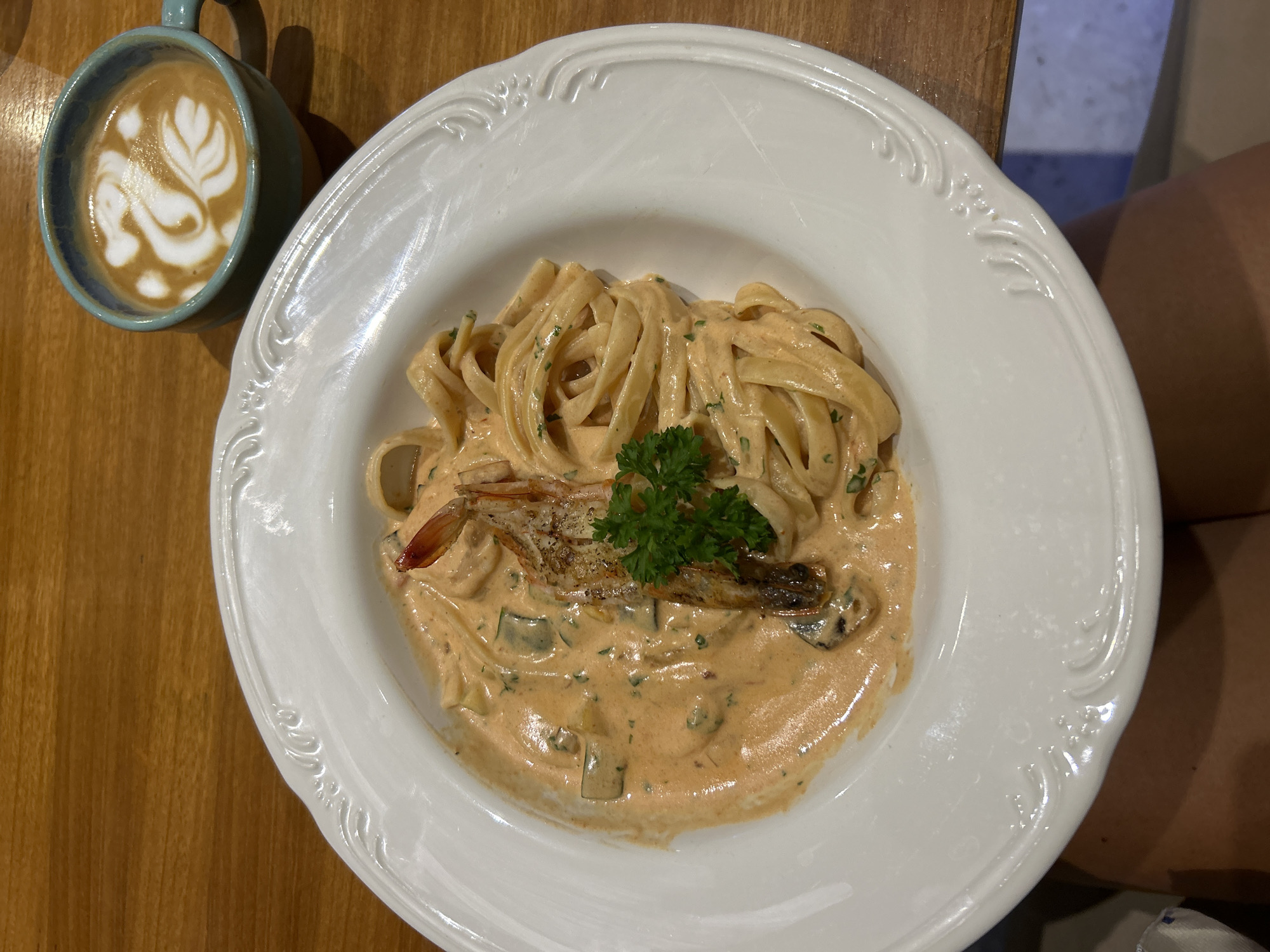 A top-down view captures a meal of creamy shrimp pasta and a latte served on a light brown wooden table, likely within a cafe or restaurant setting in Kuta Selatan, Indonesia. The central focus is a wide, white ceramic bowl with an ornate, embossed pattern along its rim. Inside, flat pasta noodles, possibly fettuccine, are generously coated in a reddish-orange creamy sauce containing chopped green herbs and small vegetable pieces. A cooked shrimp, appearing grilled with its head and tail intact, rests on top of the pasta, garnished with a fresh sprig of parsley. To the left, a teal-colored ceramic mug holds a latte with intricate white latte art, depicting a flowing, leafy or heart-like design. A person's knee is subtly visible in the bottom right corner, suggesting someone is seated at the table. The lighting indicates an indoor setting. No discernible text is present.