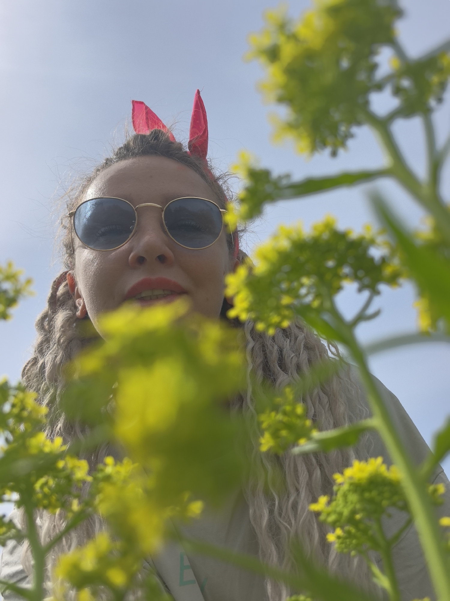 A low-angle close-up photograph depicts a woman wearing round, gold-framed sunglasses and a red headscarf fashioned into bunny ears. Her long, braided light-blonde hair frames her face. She has fair skin and wears red lipstick. Yellow flowering plants with green stems and leaves partially obscure the view of the woman and the light blue sky. A small portion of a light-colored garment with a blue-green design is visible below the woman’s chin. The photograph was probably taken outdoors in a sunny environment, but the precise location within Noviy, Russia, is impossible to ascertain.