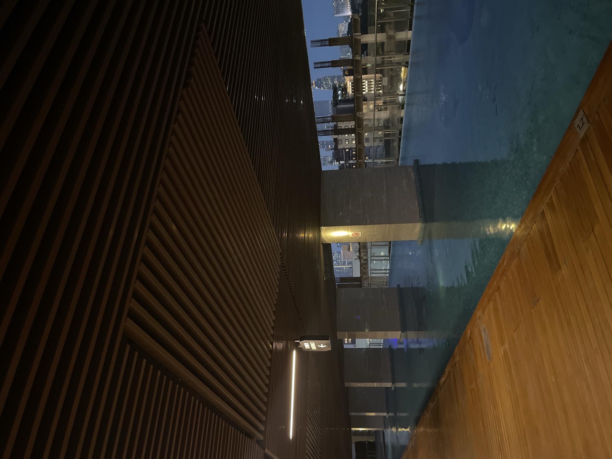 The image depicts an outdoor swimming pool area at night, likely on a rooftop or elevated level, overlooking a city skyline. The pool itself is a deep blue, reflecting the ambient lights from the surrounding buildings and the interior lighting of the pool area. A wooden deck with a warm, polished finish borders the pool on the left side of the frame. Above, a slatted wooden ceiling structure extends across the scene, providing a covered walkway or lounge area.
Visible in the mid-ground are several large, rectangular pillars that support the ceiling structure and seem to demarcate different sections of the pool area. One pillar on the left has a directional sign pointing left with text reading "TOWER EMERALD." In the distance, through the open sides of the pool area, a cityscape is visible with numerous illuminated buildings of varying heights, suggesting an urban setting. The sky above is dark blue, indicative of twilight or early night.
There are no people visible in the image. The overall impression is one of a modern, upscale amenity within a city, offering a serene escape with a view. The scene suggests a location in Central Jakarta, Indonesia, given the architectural style and density of the surrounding urban landscape, which is characteristic of that area. A depth marker "12" is visible on the edge of the pool.