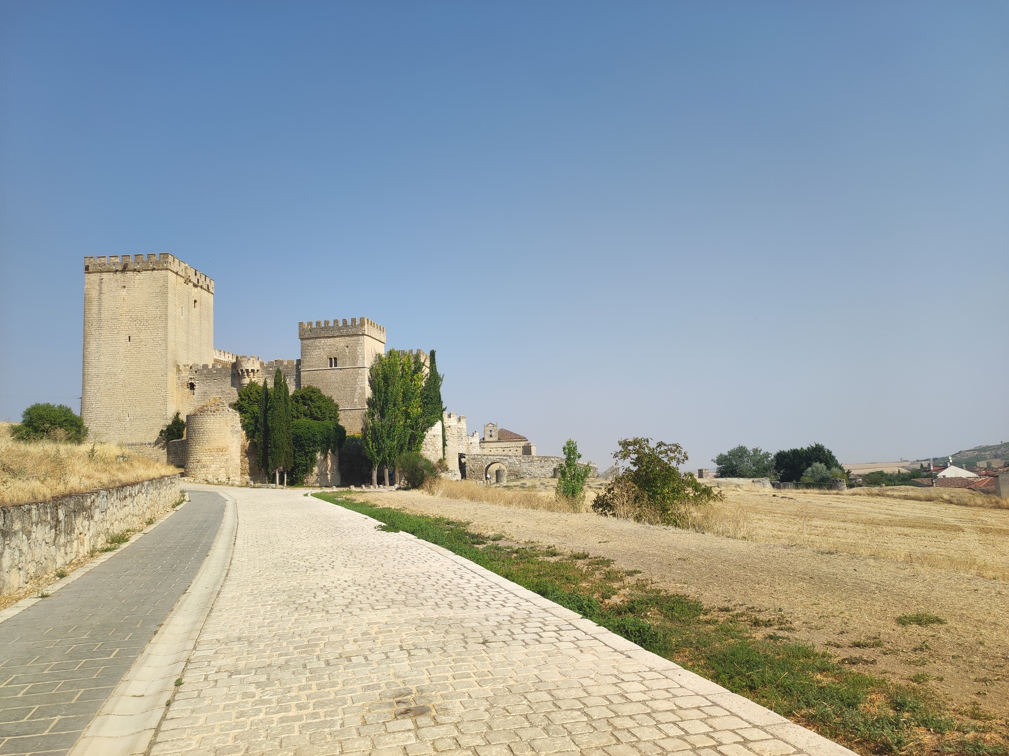 The image captures a view of the Castillo de Ampudia in Ampudia, Spain. In the foreground, a paved walkway with a stone wall to its left curves to the right, leading towards the castle. The castle, constructed from light-colored stone, features crenellated towers and walls, with a prominent keep on the left. To the right of the castle, the landscape transitions to a dry, grassy field under a clear blue sky. Distant structures, including buildings with red roofs, are visible on a gentle slope in the background, suggesting the proximity of a village or town. The scene is bathed in sunlight, indicating daytime. No people are present in the image.