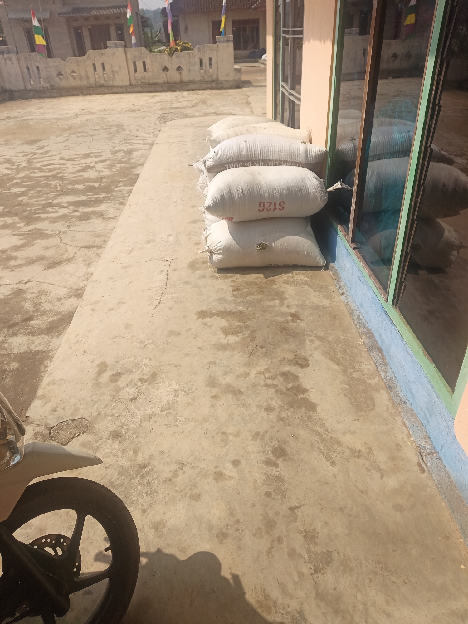 In a sun-drenched outdoor setting, likely a yard or patio, several large white sacks are stacked neatly. The sacks are printed with text, including "S12G" and what appears to be "JUAL" (Indonesian for "sell") on one of them. To the right, a building with large windows and a peach-colored facade is visible, with a blue-painted base. The ground is paved with concrete, showing signs of wear and tear with cracks and water stains. In the lower left corner, a portion of a white motorcycle, including its wheel and fender, is captured in the frame, casting a shadow. In the background, a low concrete wall with decorative cutouts separates the paved area from a building with a tiled roof, adorned with Indonesian flags. The overall scene suggests a storage area or a marketplace in a residential or commercial setting in Terbanggi Marga, Indonesia.
