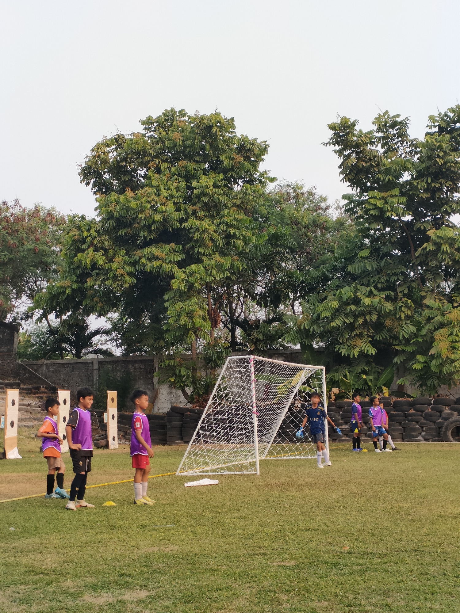 In a grassy outdoor field in Banguntapan, Indonesia, a group of young boys are participating in a soccer practice. A portable soccer goal with a white net stands on the right side of the frame. Several boys, wearing athletic uniforms consisting of purple vests over t-shirts and shorts, are positioned on the field. One boy, dressed in a blue jersey and shorts, stands near the goal, appearing to be a goalkeeper. The background features lush green trees and a grey wall. In the distance, there are also stacks of tires, possibly used as an obstacle or barrier. The overall scene suggests an organized sports activity for children in a semi-urban environment.