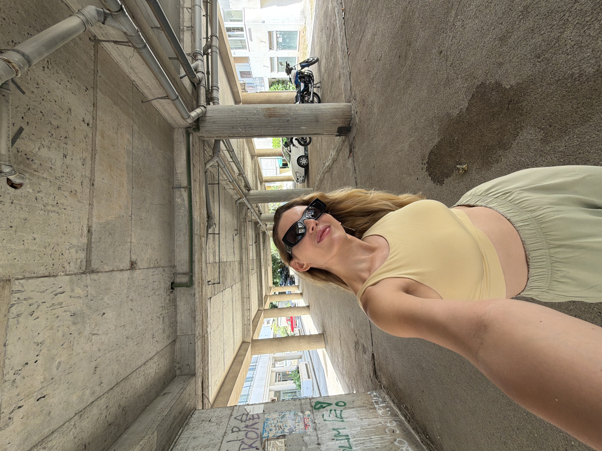 A blonde woman wearing sunglasses, a yellow crop top, and light green pants stands in a concrete underpass in Budva, Montenegro. She is taking a selfie with her left arm extended. The underpass has thick concrete pillars and a rough concrete ceiling with visible pipes. Graffiti is present on the concrete wall to the left. In the background, there are buildings, a red car, a white car, and a blue motorcycle parked on the pavement. A dark wet stain is visible on the ground in the mid-right of the image. The overall scene suggests an urban outdoor environment.