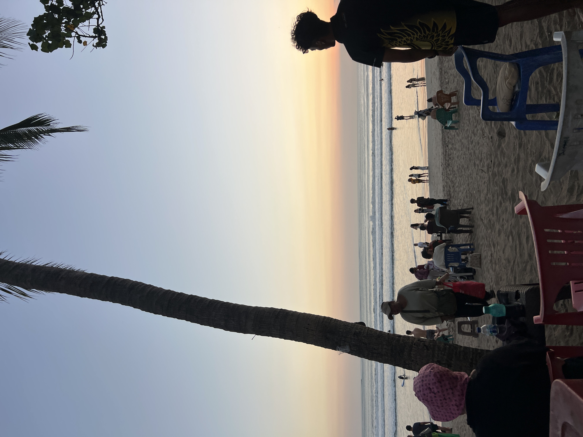 This is a photograph taken at Kuta Beach, Bali, Indonesia, during a sunset. In the foreground, several people are seated at tables on the sand. A large palm tree is positioned on the left side of the frame, its trunk textured and dark. The middle ground of the image shows a beach populated with individuals walking, sitting on chairs, and interacting. The ocean is visible with gentle waves rolling in, and several people are in the water, some appearing to be surfing. The sky exhibits a gradient of colors, transitioning from a light blue at the top to soft hues of orange, pink, and yellow near the horizon, indicative of a sunset. The overall atmosphere is relaxed and communal, typical of a popular beach destination.