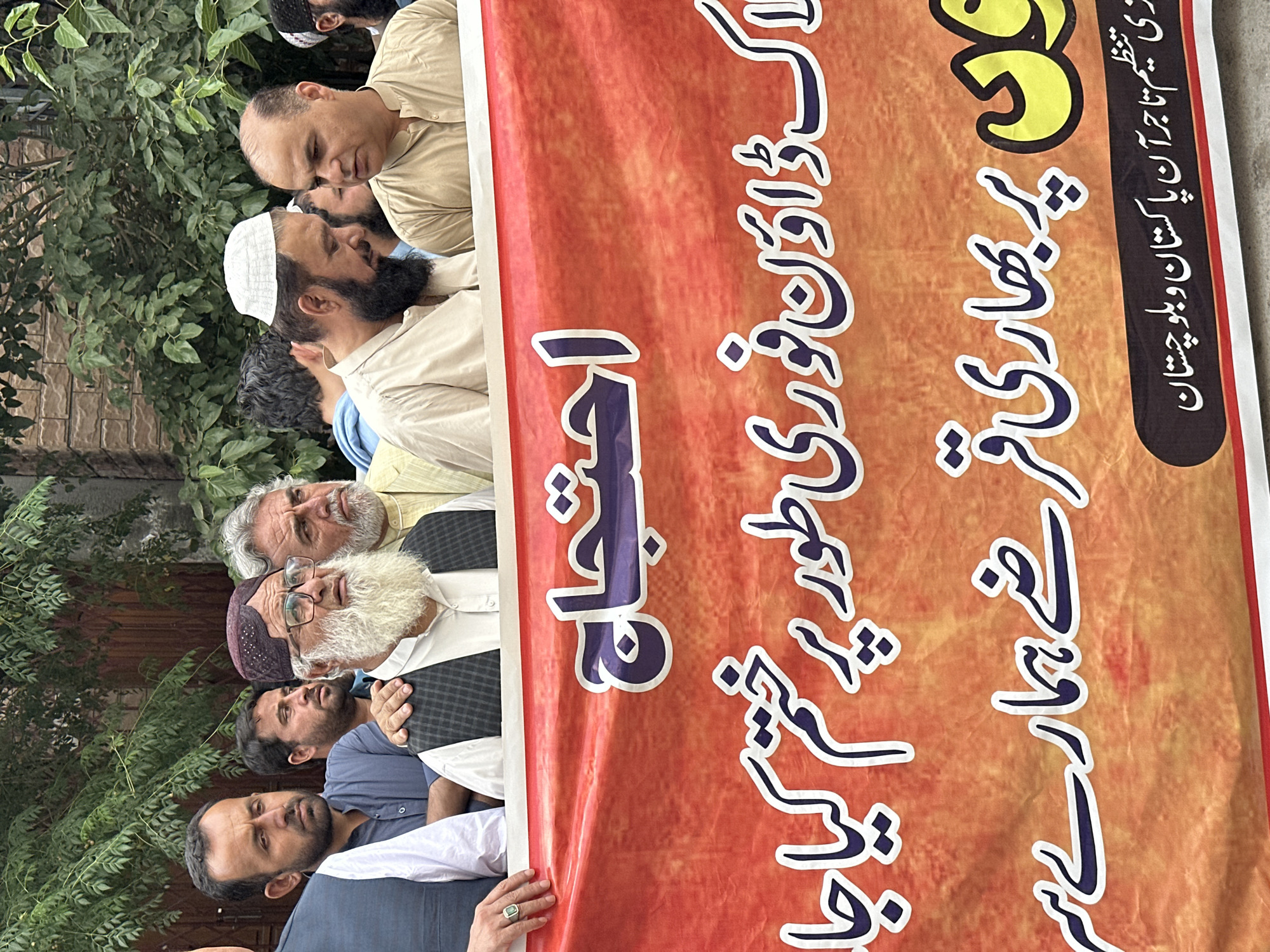 The image shows a group of men holding a banner with Urdu text. The banner features a yellow and orange background with blue text. The Urdu text on the banner translates to "Protest", "End the lockdown immediately", "Heavy", and "Organization of Traders of Pakistan and Balochistan is ours". The men are standing in front of a green leafy background, possibly plants or trees. The location appears to be an outdoor setting, possibly a street in Quetta City Tehsil, Pakistan.