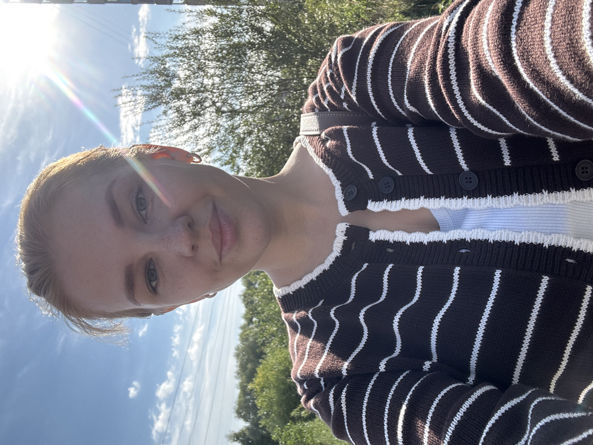 A young Caucasian woman with blonde hair pulled back is taking a selfie outdoors in Rusanovo, Russia. She is wearing a brown cardigan with white stripes over a white top. The sun is shining brightly, creating a lens flare effect and casting shadows. Trees and power lines are visible in the background under a blue sky with scattered clouds.