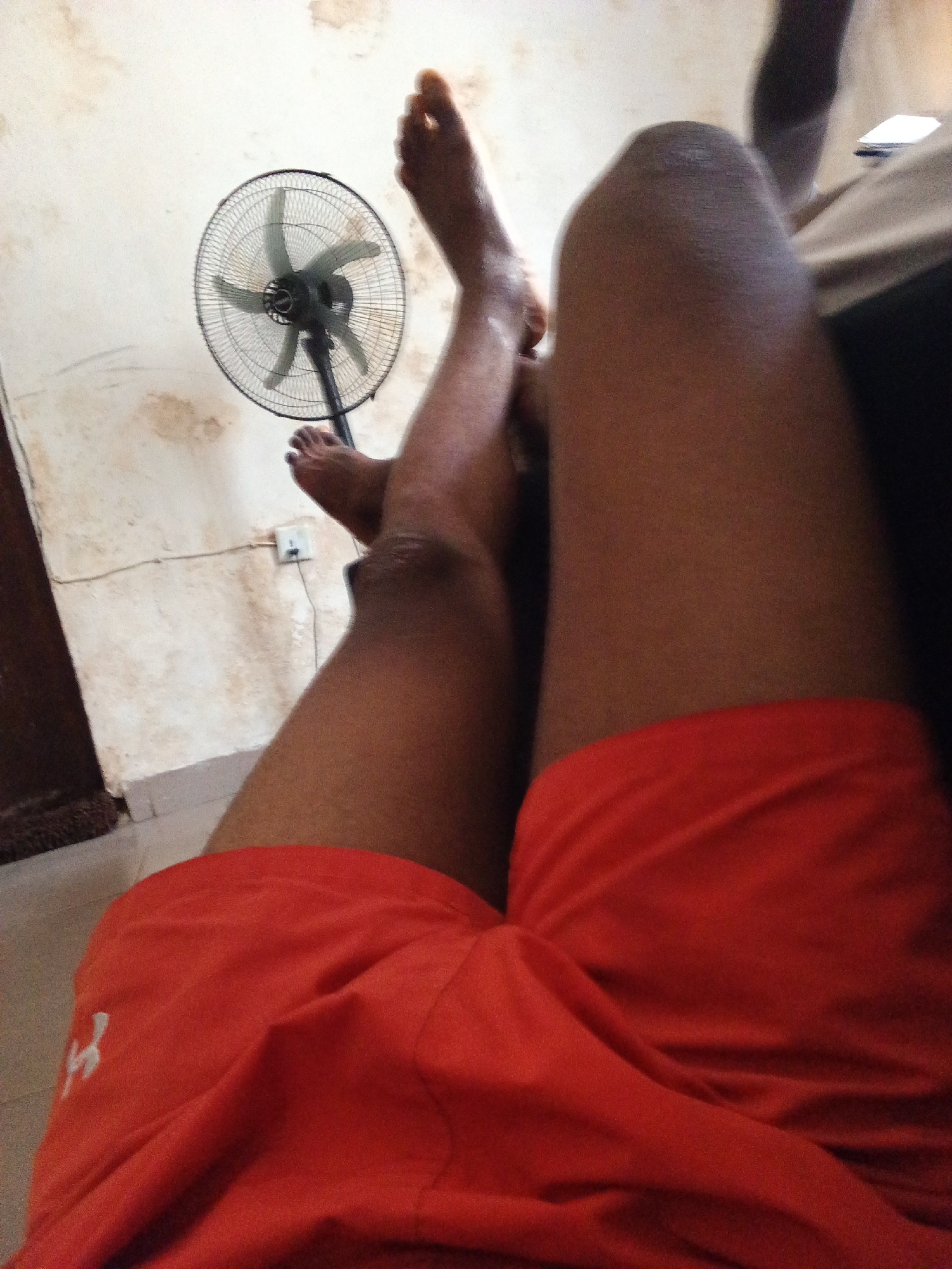 In Ado, Nigeria, a close-up shot shows the legs of a person wearing orange shorts. The shorts have a white logo on the left thigh. The person's legs are bare, and one foot is visible, pointing upwards. Behind the legs, a black oscillating fan is positioned on the floor. The wall behind the fan is stained and off-white, with an electrical outlet visible on it. The scene appears to be indoors, possibly a living space.