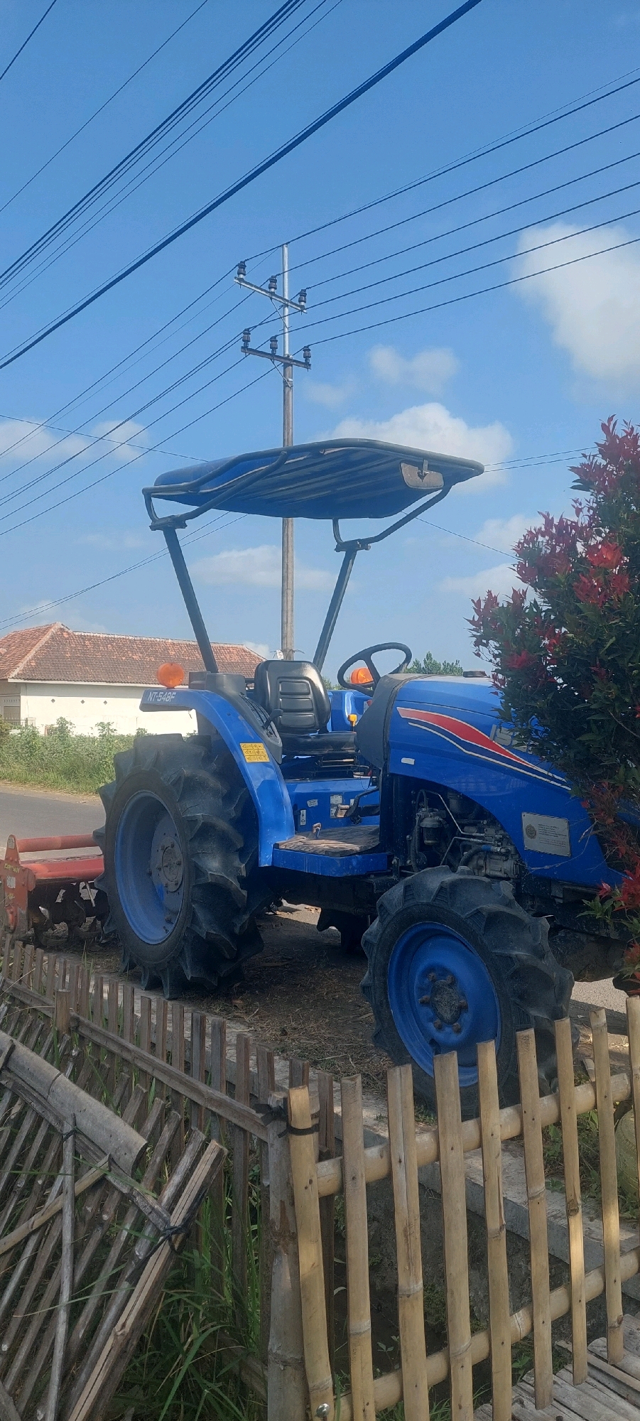 The image shows a blue tractor parked on the side of a road in Sumbersuko, Indonesia. The tractor has a canopy, a driver's seat, and large wheels. A red rotary tiller is attached to the rear of the tractor. A bamboo fence is in the foreground, and a house with a tiled roof is visible in the background. Power lines and a utility pole are also present in the scene. The sky is blue with some clouds. The overall scene depicts a rural or semi-rural setting in Indonesia.