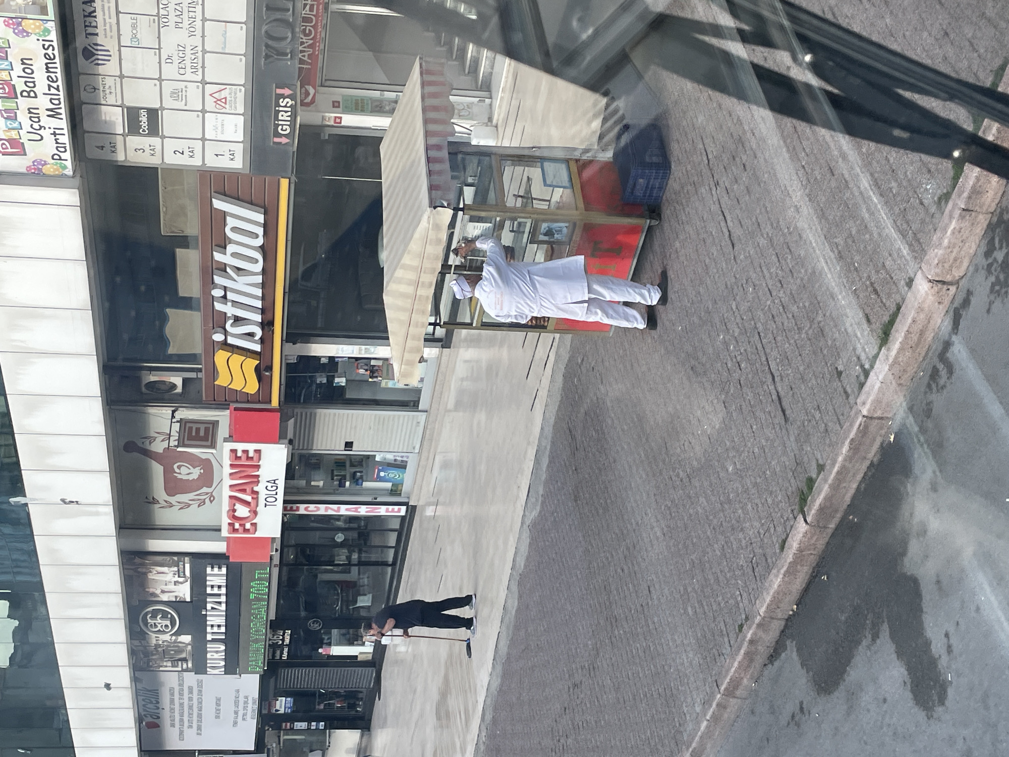 A street scene in Kadıköy, Turkey, shows two men. One man, wearing a dark shirt and pants, is sweeping the sidewalk with a broom. Another man, dressed in white with a cap and white coat, is cleaning the glass of a food cart. The food cart has a red base and a striped awning. Behind the men, various storefronts are visible, including signs for "Arçelik," "KURU TEMİZLEME," "ECZANE TOLGA," and "istikbal." The ground is paved with textured concrete, and a wet patch is visible on the lower left. The background reveals a modern building facade.