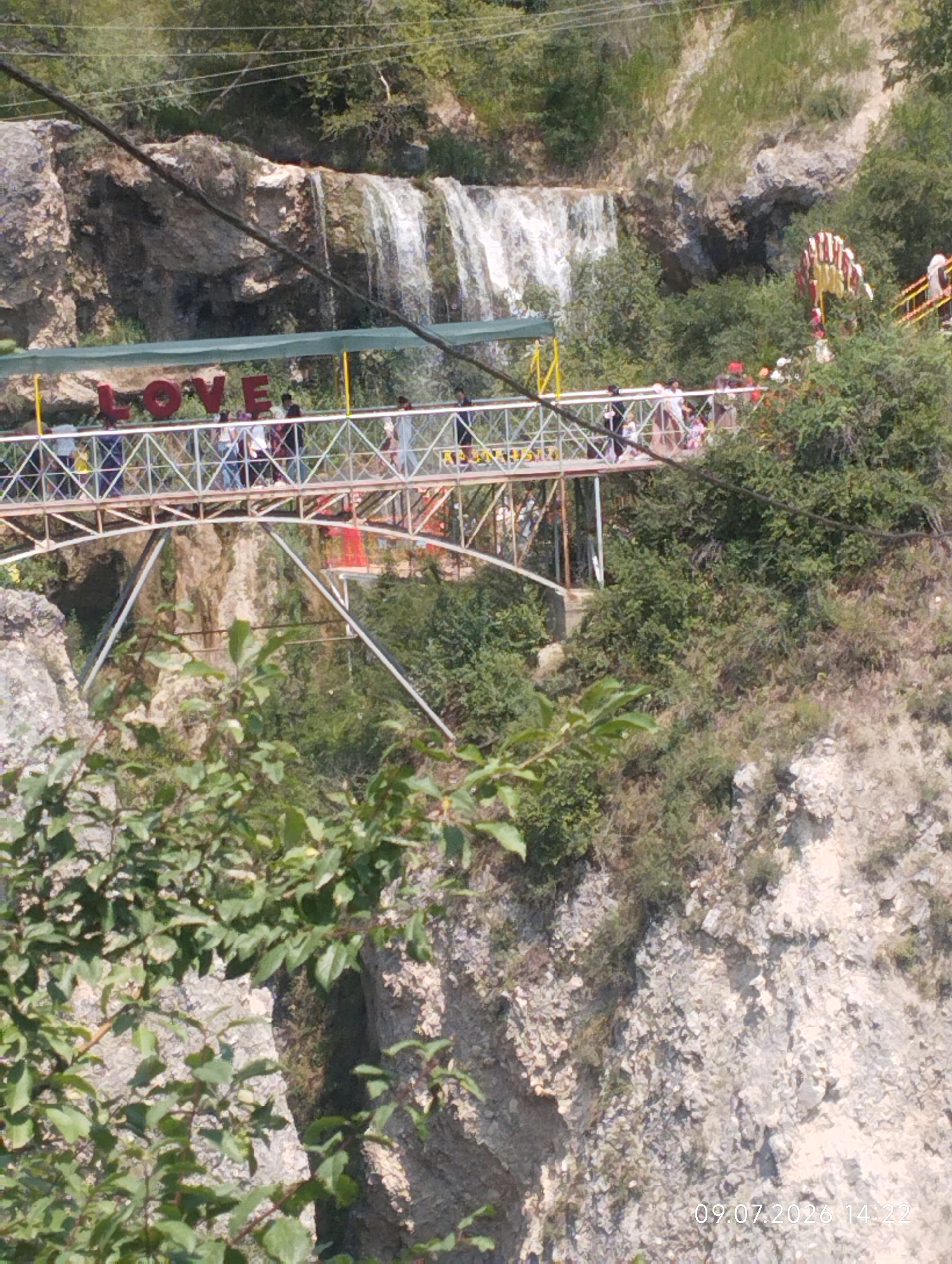 The image shows a scenic view of a waterfall in Arstanbap, Kyrgyzstan. A pedestrian bridge spans across the gorge, with the word "LOVE" written in large red letters above it. Several people are visible on the bridge, some walking and others observing the waterfall. The surrounding landscape is characterized by rocky cliffs, lush green vegetation, and trees. In the background, a portion of a fairground attraction with red and white colors can be seen. The water from the waterfall cascades down the rocks, creating a misty spray. The image is dated 09.07.2026 at 14:22.