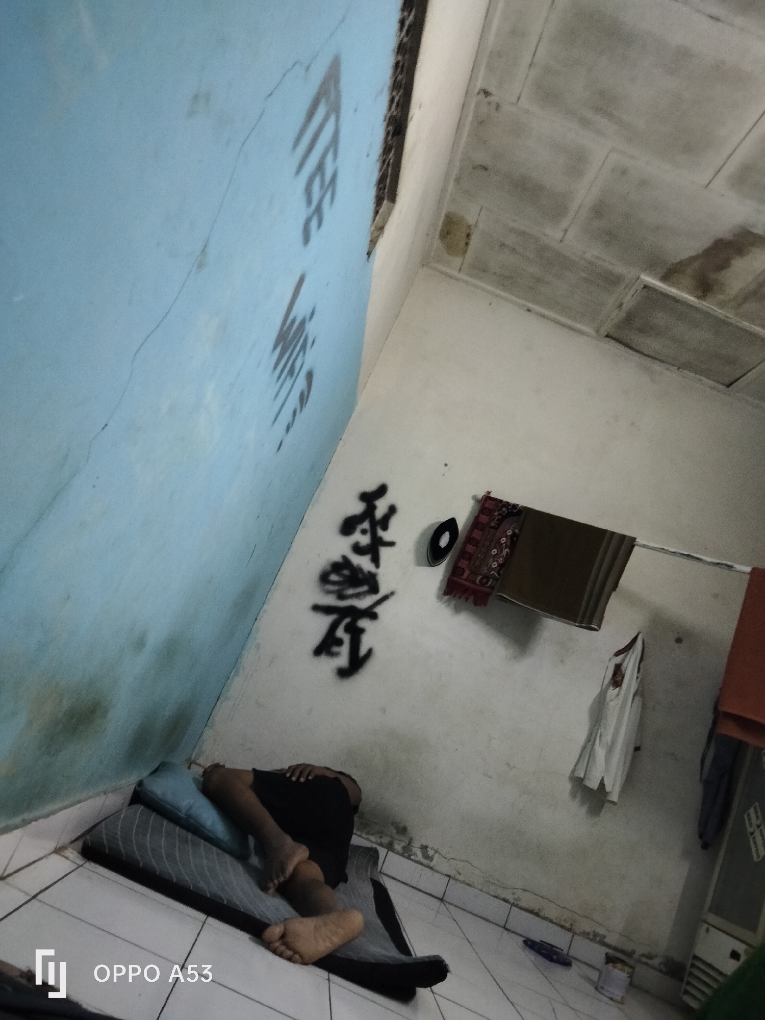 The image depicts a dimly lit room with light blue walls and a white ceiling. Graffiti, appearing to be in black paint, is present on the walls. One set of graffiti, on the left wall, consists of several vertical lines. Another set, on the right wall, comprises two characters in what seems to be an East Asian script.
In the foreground, a person is lying on their side on a mat and a pillow, wearing black shorts and appearing to be asleep. Their legs and feet are visible.
Various objects are scattered around the room. On the right wall, a dark circular object, possibly a hat, is hanging next to a folded piece of fabric, possibly a sarong or prayer mat, with intricate patterns. A white garment is also hanging on the wall. Towards the bottom right, part of a white appliance, potentially an air conditioner or cabinet, is visible, along with a can on the floor. The floor is tiled with a grid pattern.
The lighting suggests it is nighttime or the room is poorly illuminated. The overall impression is that of a simple, somewhat cluttered living space. The image's timestamp indicates it was taken in Denpasar, Indonesia.