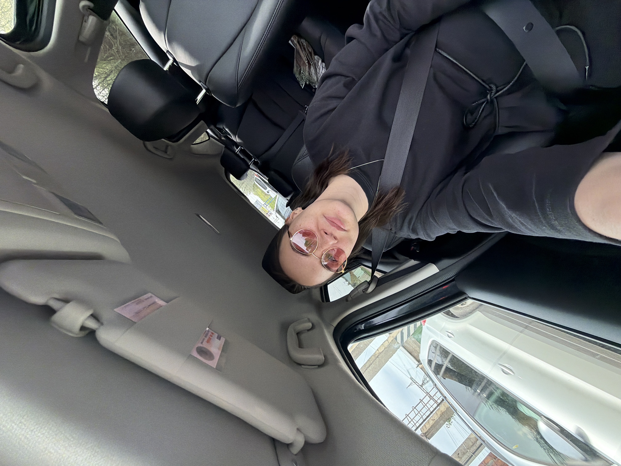 A woman wearing rose-tinted sunglasses and a black outfit is inside a car. She is wearing a seatbelt. The car interior includes grey trim and black leather seats. The sun visor has papers tucked into it. Through the window, another car and urban elements are visible. The location is Chaikovsky, Russia.