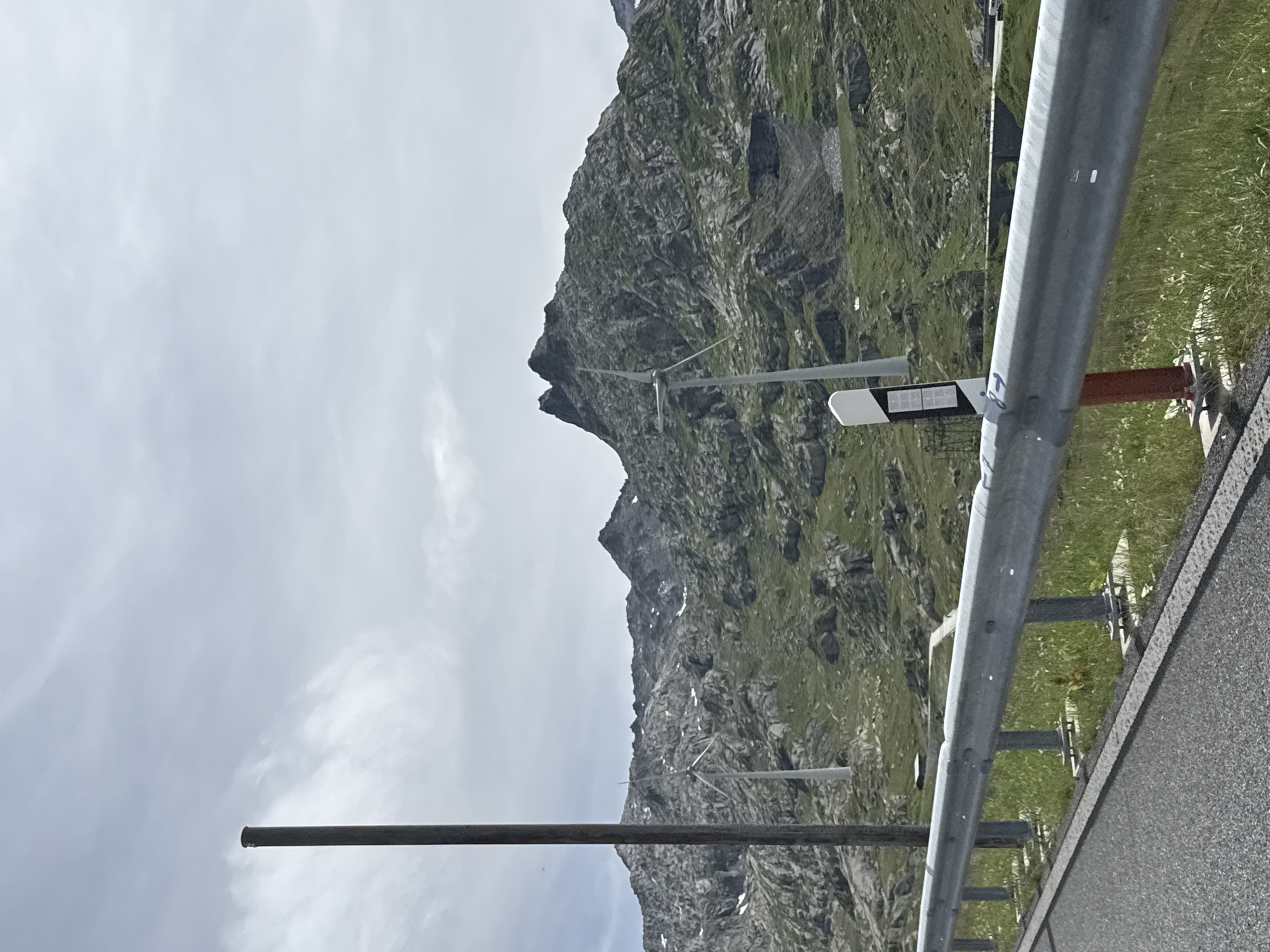 This image depicts a mountain scene, likely in Airolo, Switzerland, given the context. The foreground is dominated by a road barrier made of metal poles and a silver rail. To the left of the barrier, a long, dark metal pole extends upwards from the bottom left corner of the frame and disappears near the top. The background features a steep, rocky mountain covered in patches of green grass and small patches of snow. The sky is overcast with soft white clouds, suggesting a cool or temperate day.
Several pieces of infrastructure are visible along the roadside and on the mountain. A white and grey object with a dark rectangular panel is mounted on a pole near the road barrier. Further up the mountain, the faint outlines of what appear to be power lines or a ski lift system can be discerned. The overall impression is one of a remote, natural landscape with some human intervention for infrastructure. There is no visible text in the image. The activity suggested is simply the presence of a road, likely a scenic route, through the mountains. The weather appears to be overcast, and the time of day is indeterminate due to the lack of distinct shadows.