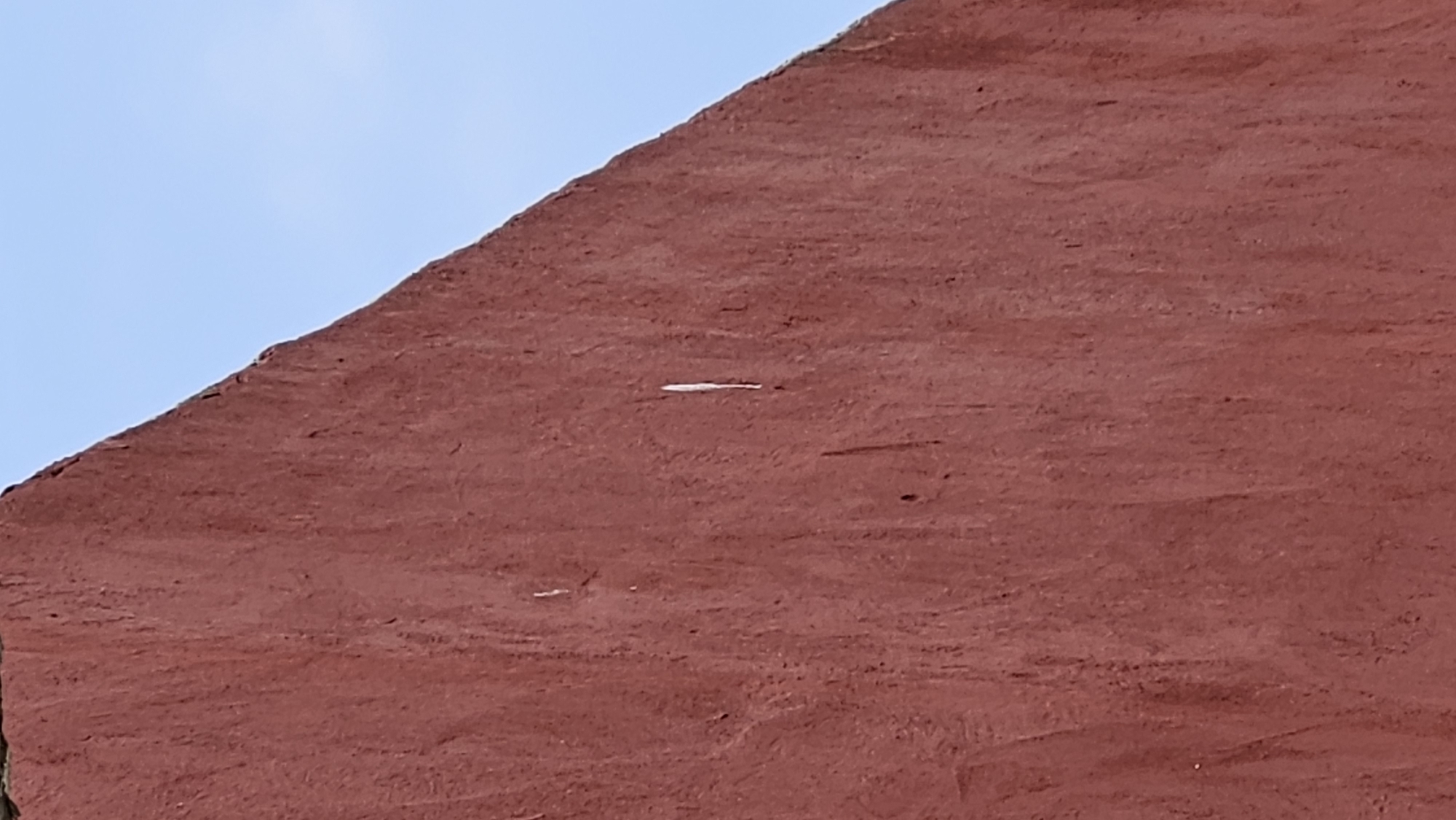 The image shows a close-up of a rough, textured, reddish-brown wall against a clear blue sky. The wall takes up the majority of the frame and has a slightly uneven, weathered appearance with vertical striations. A thin, vertical white mark is visible on the wall, slightly off-center and to the right. The top edge of the wall forms a triangular shape against the sky. The scene is a detail of an exterior wall.
