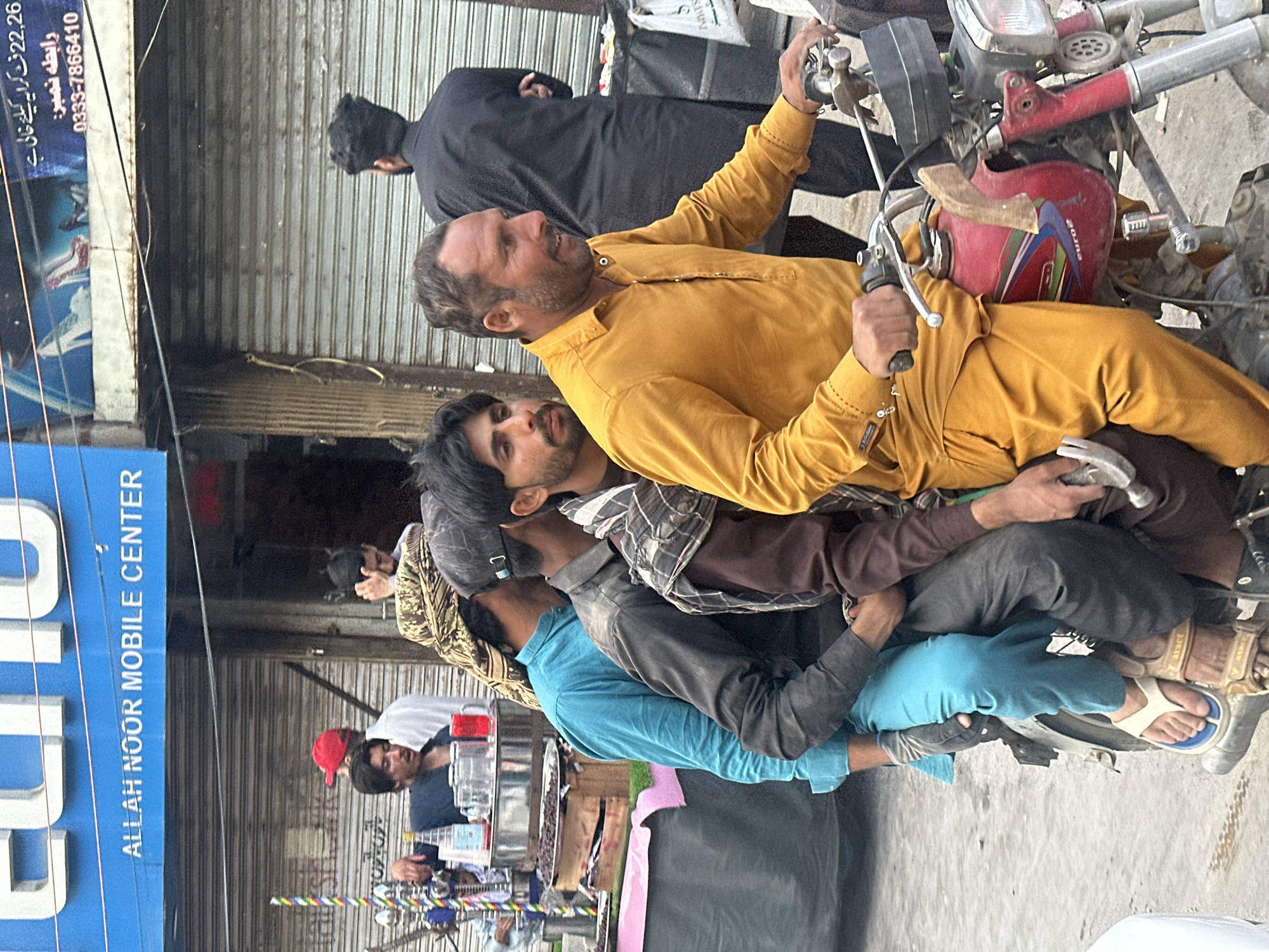 Here is a description of the image:
**Scene:** A daytime street scene in what is likely a commercial area of Quetta City Tehsil, Pakistan. The image is taken at a slight angle, looking downward.
**People:** The main subjects are several men riding a motorbike. The driver is wearing a yellow shirt and is in the front, operating the vehicle. Behind him is at least one other man, wearing a darker brown shirt. Further back is a man wearing a blue shirt and head covering. Several other people can be seen standing or walking in the background, including one person wearing a red cap.
**Objects:** A central element is a red motorbike, which appears to be a well-used model. A blue sign for "ALLAH NOOR MOBILE CENTER" is visible on the left side of the frame, indicating the presence of a mobile phone shop. Other elements include a stand selling snacks/food products, various buildings with closed metal roller shutters, and utility poles with wires.
**Actions/Interactions:** The primary action is the men riding on the motorbike. It appears as though multiple people are riding on a vehicle designed for fewer passengers, indicating overcrowding. There is pedestrian traffic in the background, and some individuals seem to be engaged in commerce.
**Environment:** The area seems to be a busy street with both pedestrian and vehicle traffic. The buildings are simple, commercial structures with metal shutters. The lighting is natural daylight.