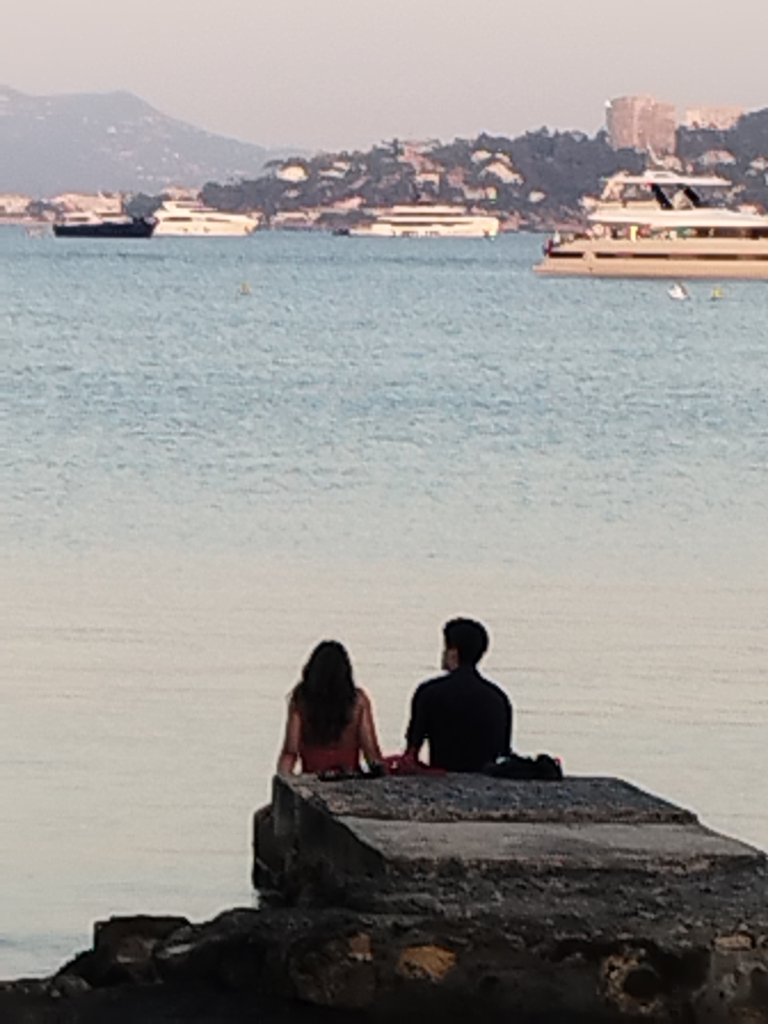 The image depicts a serene waterside scene with two people sitting on a concrete structure facing away from the camera. The couple appears to be a man and a woman, with the woman on the left wearing a red garment and the man on the right wearing a dark, long-sleeved shirt. They are seated close together, looking out at the water.
In the background, several boats are visible on the calm sea. One large, white yacht is prominent in the middle ground, and another dark vessel is further to the left. Beyond the water, a coastline is visible with buildings nestled amongst trees on a hillside. The sky is hazy, suggesting it may be dawn or dusk, or possibly a slightly overcast day.
The overall setting evokes a peaceful and romantic ambiance, likely in a coastal city. Given the context of Antibes, France, this scene could be captured from a vantage point overlooking the Baie des Anges or near the Port Vauban, popular areas for yachts and seaside relaxation. The architecture and the type of boats visible are consistent with the French Riviera.