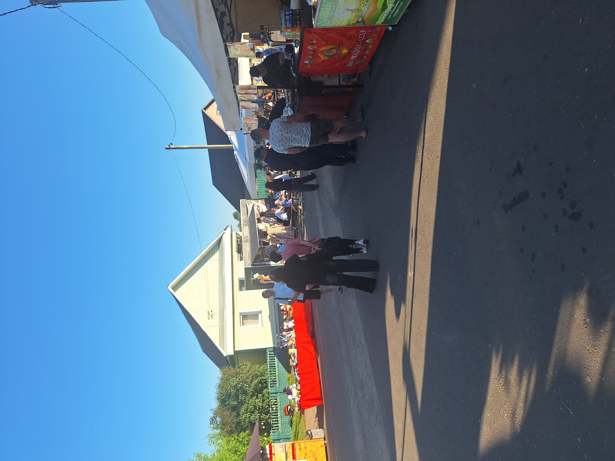 The image depicts an outdoor market scene on a sunny day, likely in Цімкавіцкі сельскі Савет, Belarus. In the foreground, the asphalt road is partially covered by the shadows of trees and structures. Several people are present, browsing and interacting at market stalls.
On the left side of the image, a yellow food stand is visible, with a red table displaying various items, possibly crafts or goods, behind it. A group of people are gathered around this area, with some appearing to be vendors and others customers. Further back, a light-colored residential house with a peaked roof is situated.
Towards the right side of the image, several white tents or canopies are set up, indicating more market stalls. One stall appears to be selling food, with a person behind a counter and bottles of beverages visible. Another stall features a red banner with Cyrillic lettering and imagery. People are seen standing and walking between these stalls. The background reveals more buildings and trees, characteristic of a rural or semi-rural setting. The sky is clear and blue, suggesting fair weather conditions.