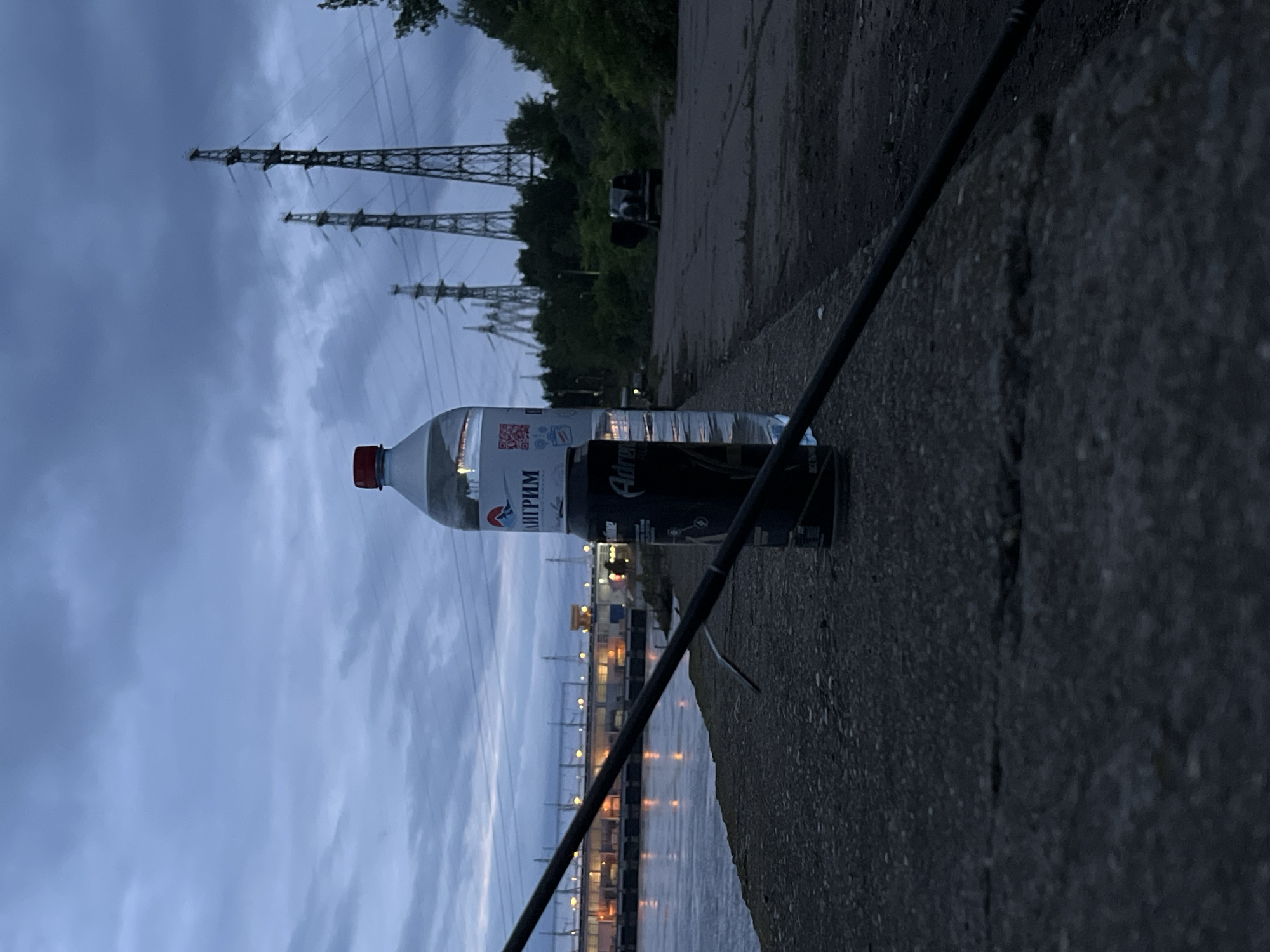 The image depicts a twilight or early night scene in Noviy, Russia, characterized by an industrial waterfront environment. In the foreground, a dark, thin fishing rod rests diagonally on a textured, dark gray concrete ledge. Positioned on this ledge are a clear plastic water bottle with a red cap, displaying the brand "ПИЛИГРИМ" (Piligrim) and a QR code on its label, and a partially visible black energy drink can, identifying as "Adrenalin Rush."
To the left of the frame, a body of water, likely a river or canal, reflects numerous yellow and orange lights from an illuminated industrial complex or dam structure on the opposite bank. This structure features multiple levels and visible conduits or railings. To the right of the frame, beyond the concrete ledge, an unpaved or worn asphalt path leads into the distance, flanked by dense green vegetation. A dark-colored vehicle, possibly an SUV or hatchback, is partially visible further down the path, with its rear hatch open. The background is dominated by a series of tall, metal electricity transmission towers (pylons) supporting power lines, silhouetted against a cloudy, dark blue-gray sky. No individuals are present in the scene.