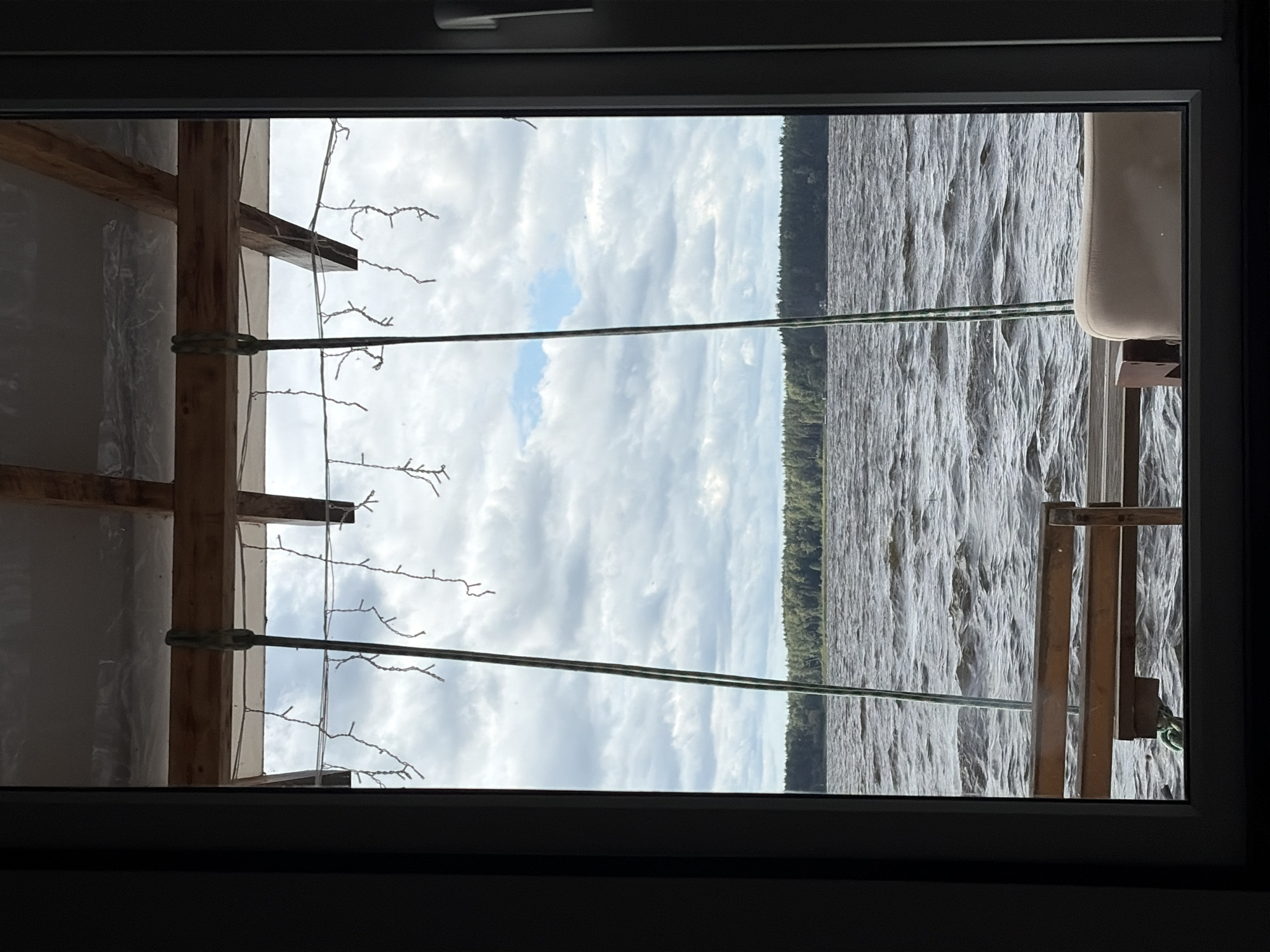 The image shows a view through a window overlooking a body of water with a treeline in the distance. The sky is cloudy. In the foreground, a wooden bench is partially visible, and a light-colored cushion is on what appears to be another seat. Two green ropes hang down from an overhead structure, and string lights are draped across the ceiling. The overall scene suggests a sheltered outdoor seating area near a lake or river. While the prompt mentions Vidogoshchi, Russia, there are no specific visual cues in the image to confirm this location.