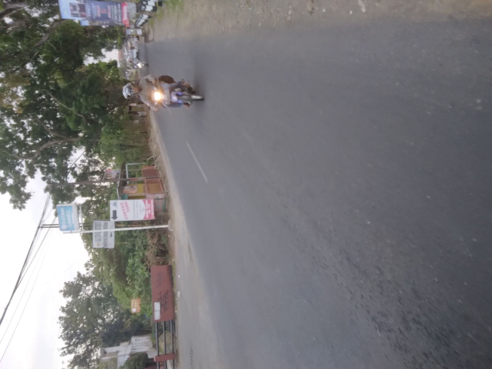 The image shows a road in Desa Pamijen, Kecamatan Sokaraja, Indonesia. A person is riding a motorcycle on the road. There are trees and buildings on the side of the road. There are also signs visible, one for "COFFEE TIME" and another for "Ice Clean Laundry". The sky is overcast.
