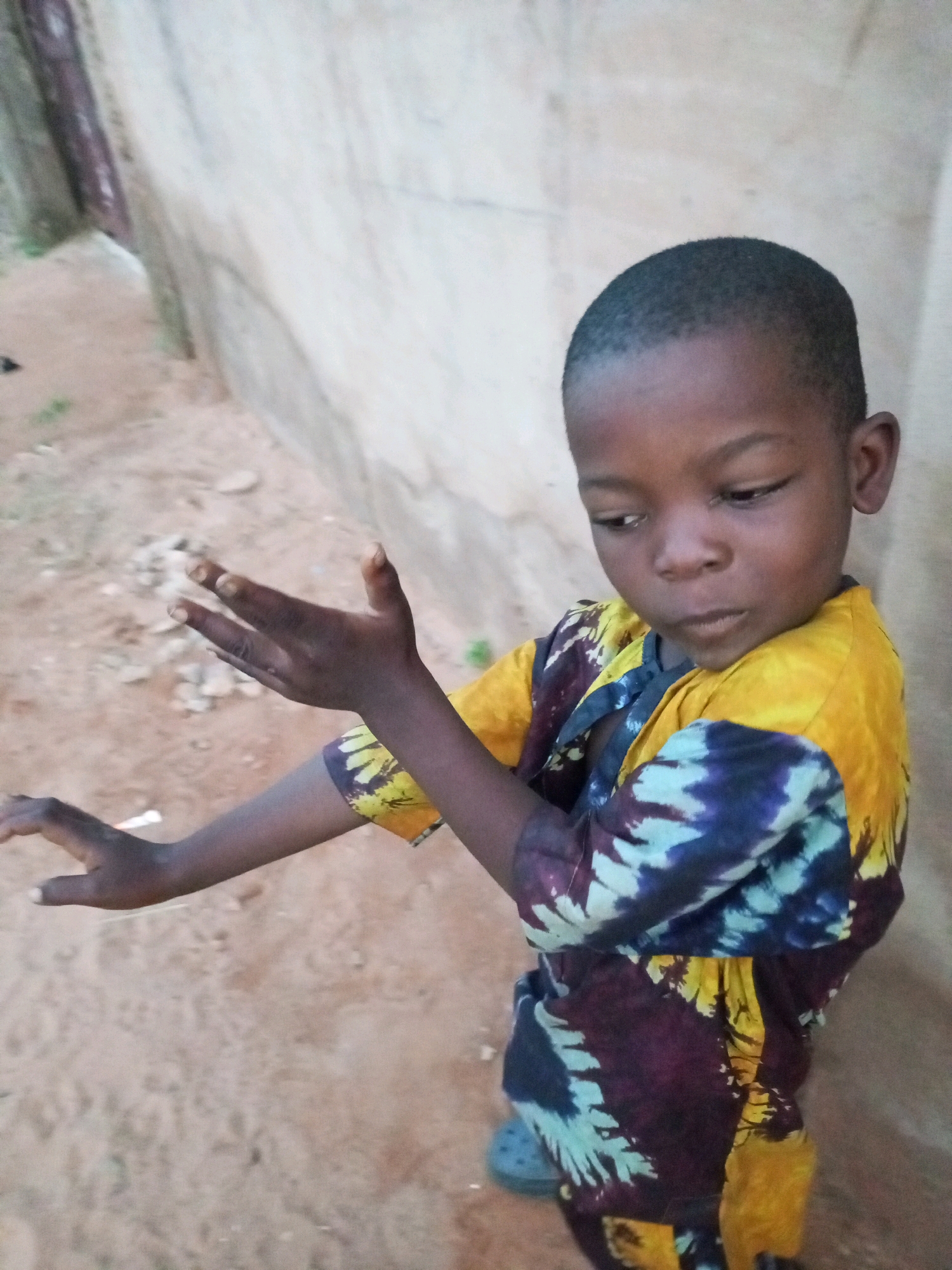 A young Black boy stands outdoors, his head and shoulders visible in the foreground. He is wearing a colorful shirt with a batik-style pattern of yellow, blue, and purple. His gaze is directed downwards and to his left, with his mouth slightly open. His left arm is extended and bent at the elbow, with his hand open and facing upwards, fingers spread. His right arm is bent at the elbow and held closer to his body, with his hand resting on his chest.
The background consists of a textured, light-colored wall to the boy's right and a sandy ground surface to his left. The wall appears to be made of concrete or a similar material and has some subtle variations in color and texture. The ground is a light brown, sandy soil with small pebbles and scattered debris, including a white object in the lower left corner. A dark, vertical post is visible along the left edge of the image, against the wall. The lighting suggests it is daytime, with diffused light illuminating the scene. The environment appears to be an outdoor setting in a possibly rural or less developed urban area. The image does not provide specific indicators of the city of Tempure, Nigeria.
