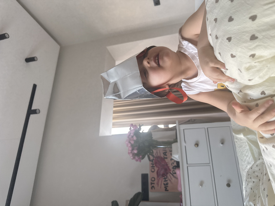 The scene depicts a young child indoors, likely in a bedroom, where the specific location within the city of Khimki, Russia, cannot be determined from the visual evidence. The child, with light skin and dark hair, is positioned on a bed and wears a white sleeveless top. A conical, reflective hat, possibly made of foil or similar material, is on their head, secured with red ribbons tied under the chin. The child's mouth is slightly open, and their gaze is directed upwards and to the right of the frame. The bed is covered with light-colored bedding patterned with small, dark heart shapes.
In the background to the left, a white dresser with multiple drawers is visible. On its surface, a vase holds a bouquet of pink roses, and a framed piece of artwork displays a textured red heart. Russian text is partially legible on the artwork, reading "ЭТО ОНА" (This is her/it) above the heart and "МЕНЯ ТАК" (me so) followed by an incomplete word starting with "РАЗЪЕ" (raz"ye, suggesting 'corrodes' or 'eats away') below. A dark green plant is partially visible to the far left. To the right of the dresser, a window is partially obscured by light-colored curtains. The ceiling is light-colored and features a black track lighting system with multiple spot fixtures. Natural light illuminates the room.