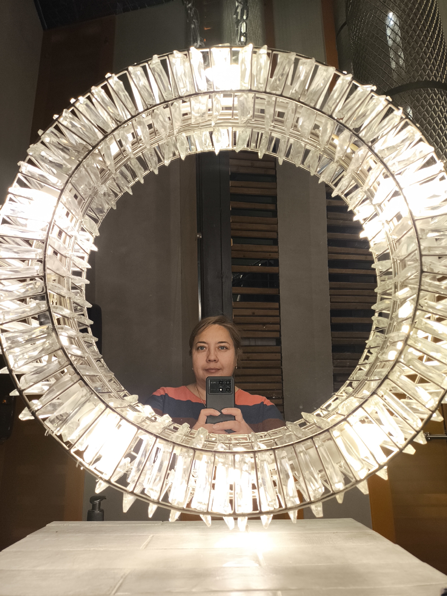 A woman is taking a selfie in a mirror with a decorative ring of clear crystals around it. The mirror appears to be located in a bathroom setting, with white tiled surfaces visible below. The woman is wearing a striped shirt and holding a smartphone. The background reflected in the mirror shows a wall with horizontal wooden slats and a textured wall covering. No information is available from the image to suggest that this image was taken in the city of Khimki, Russia.