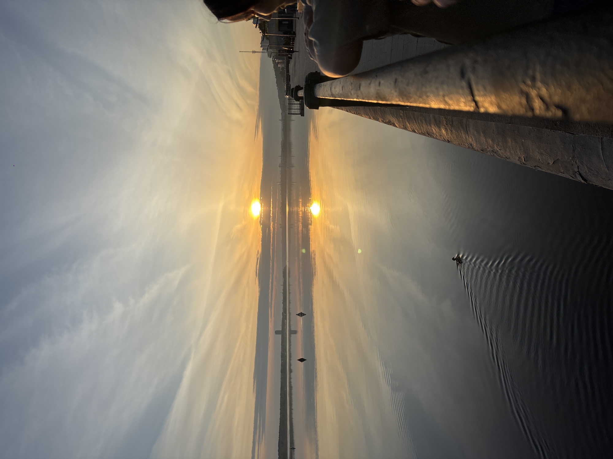 The image shows a body of water with the reflection of a sunset and the sky. A railing runs along the top right of the frame. In the background is a cityscape. A small boat is in the water. The image appears to have been taken in the city of Perm, Russia, based on the presence of the Kama river embankment.