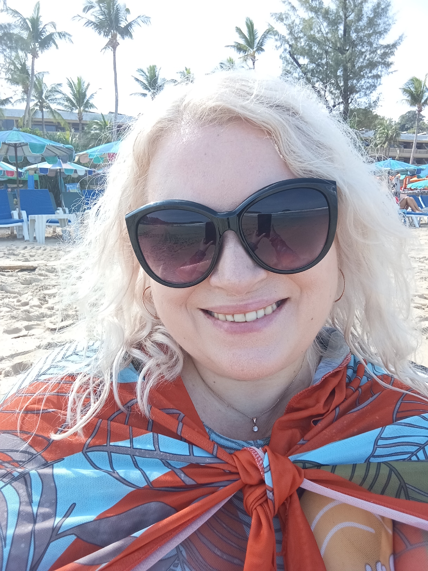 A Caucasian woman with blonde, wavy hair is taking a selfie on a beach in Karon, Thailand. She is wearing large, dark sunglasses, a thin silver necklace with a small diamond pendant, and an orange and blue patterned sarong tied around her shoulders. The background shows a sandy beach with lounge chairs and umbrellas, palm trees, and buildings. The sun is shining, and the sky is bright.