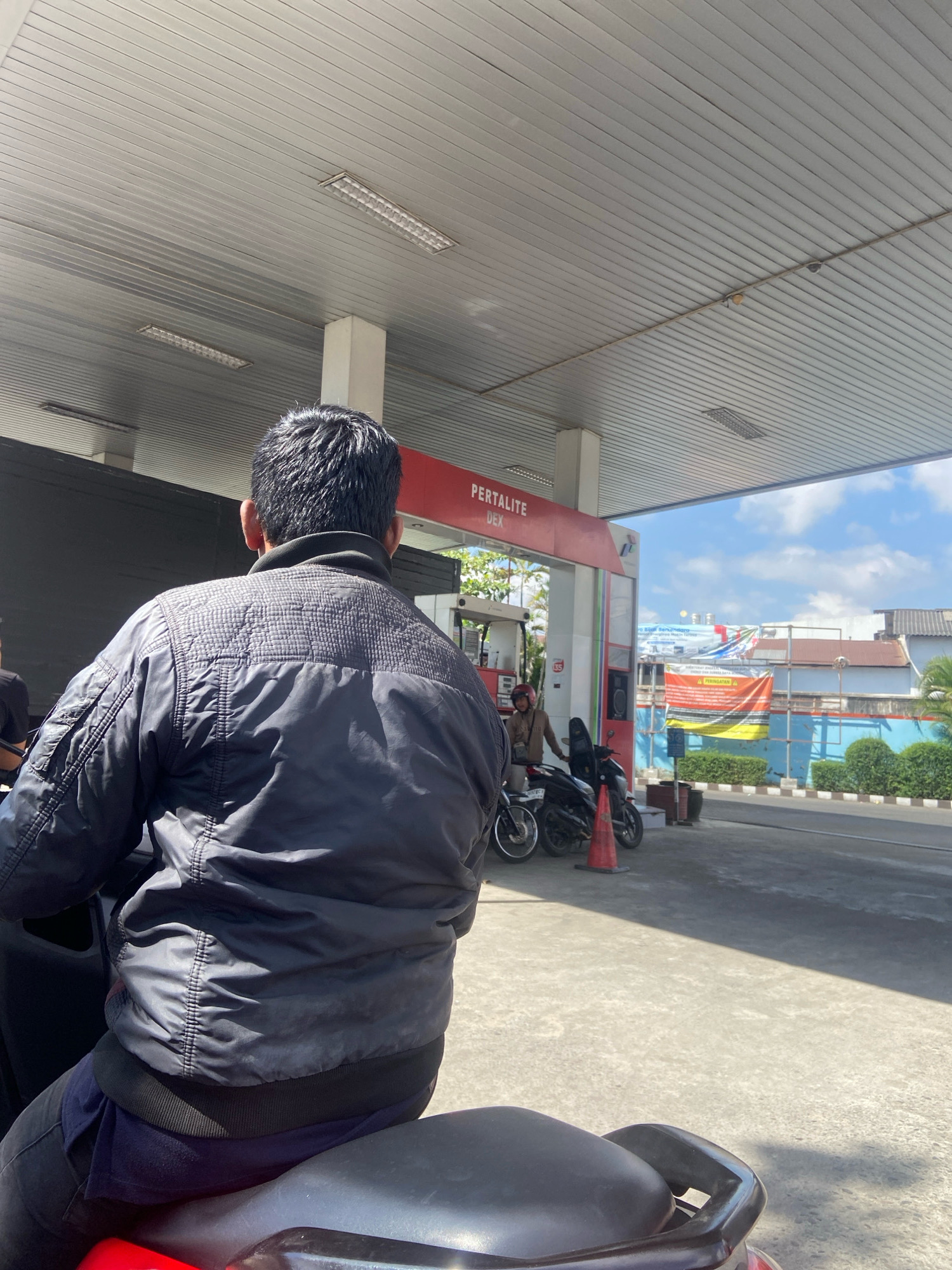 The image is an outdoor, eye-level, medium shot capturing a view from behind a person seated on a motorcycle at a Pertalite gas station in Kedungwaru, Indonesia. The person in the foreground is wearing a black puffer jacket and dark pants, facing away from the camera. The gas station features a red and white canopy with fluorescent lights. In the background, a gas pump is visible, along with several other motorcycles and two people standing near them. One person is wearing a helmet and a brown jacket, while another person's head and shoulders are visible on the far left of the frame. Banners are displayed on a building in the distance, and a clear blue sky with scattered clouds is visible above. The ground is paved concrete, with some shadows cast by the canopy.