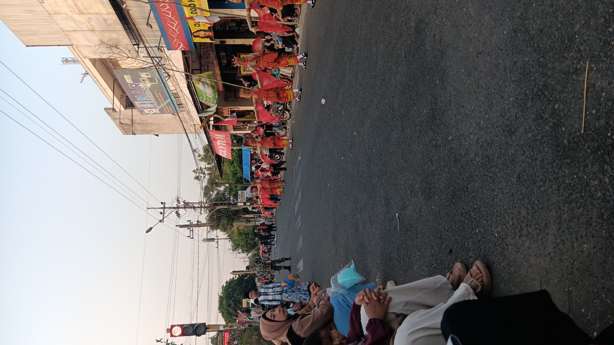 On a paved road in Tanjung Anom, Indonesia, a parade is underway. A group of performers dressed in traditional red and gold costumes, complete with headdresses and flowing red scarves, are marching in formation. They are performing synchronized movements, with their arms extended. Spectators are seated along the side of the road, watching the procession. Some individuals are standing or walking further down the street. The scene is illuminated by natural daylight, with buildings visible on the right side of the road, some bearing signs. Traffic lights and utility poles are also present along the street. The atmosphere suggests a community event or celebration.