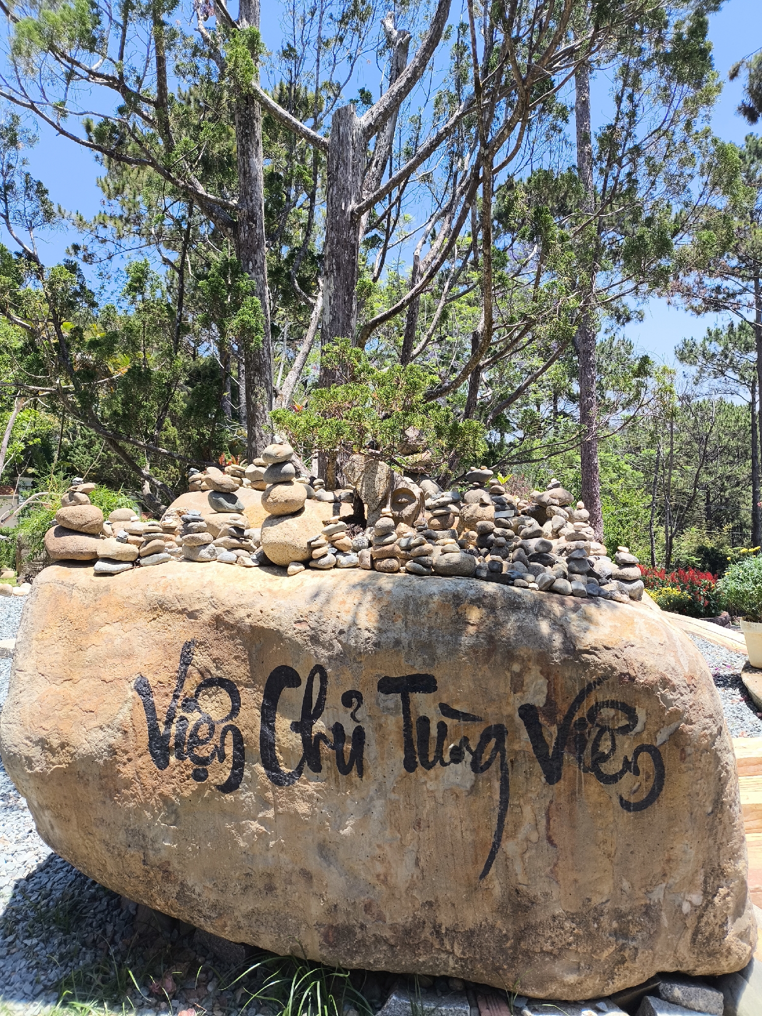 The image shows a large boulder with the words "Viện Chỉ Từng Viên" painted in black on its surface. The top of the boulder is covered with stacks of small rocks, creating numerous miniature cairns. The boulder is situated in an outdoor setting, surrounded by trees, suggesting a park or garden environment. In the background, there is a mix of evergreen trees and glimpses of other vegetation, including some flowering plants. The sky is visible in the upper portion of the image, appearing clear and blue. This rock with the writing is located in the city of Da Lat, Vietnam.