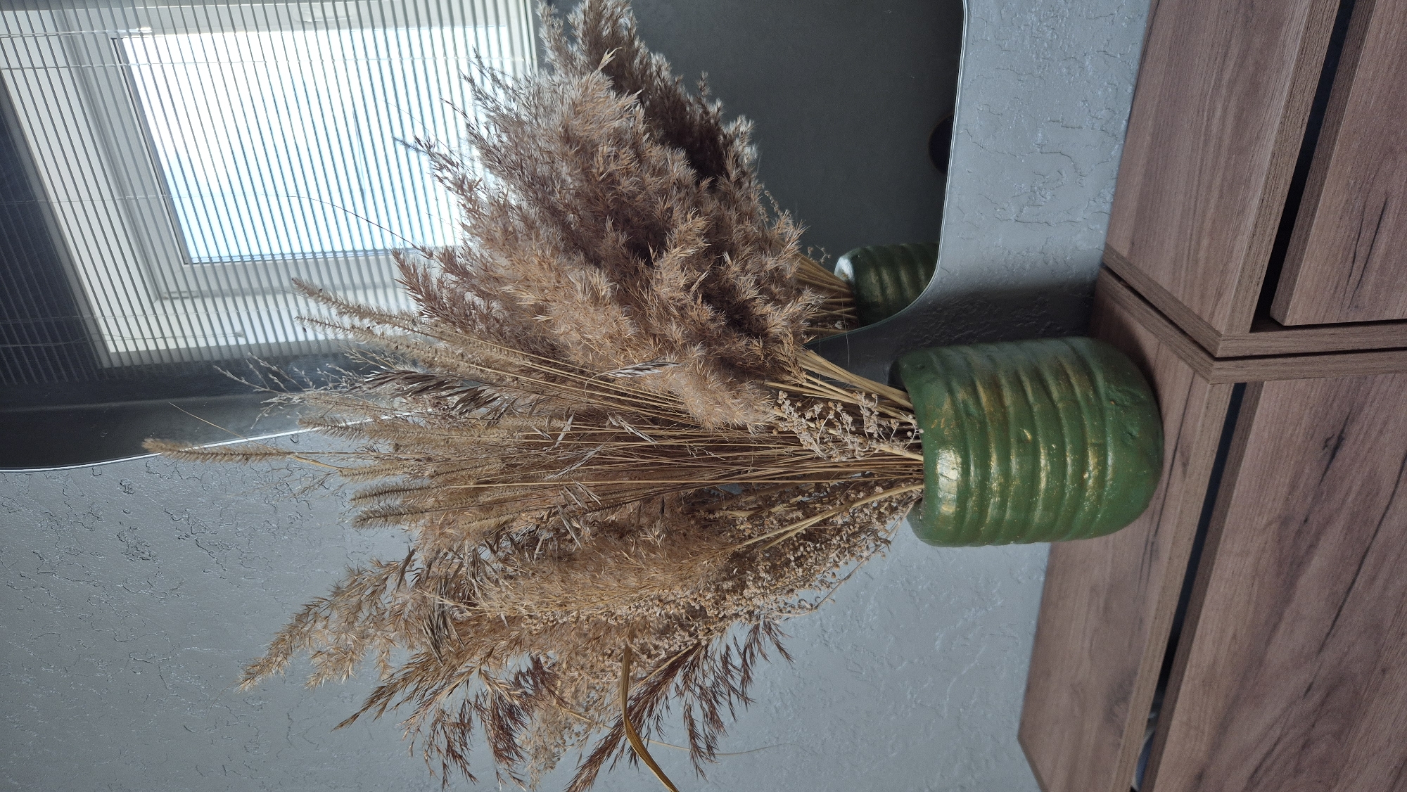 A close-up shot captures a decorative arrangement of dried grasses in a textured green vase. The vase, with its ribbed design and subtle gold accents, sits on a wooden surface that appears to be a dresser or shelf. Behind the vase, a large mirror reflects the scene, including a window with horizontal blinds that are partially open, allowing light to stream in. The wall behind the arrangement has a textured gray finish. The overall scene suggests an indoor setting, likely a home, with a focus on natural textures and a neutral color palette. There are no people or animals present in the image. The location in Pomryaskino, Russia, is not visually discernible from the provided image.